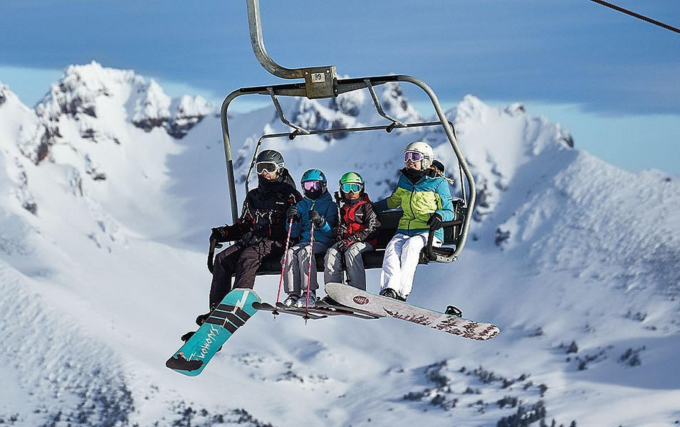 A group of skiers and snowboarders wearing Columbia Sportswear ski gear rides a chairlift with a sunny mountain range in the background.