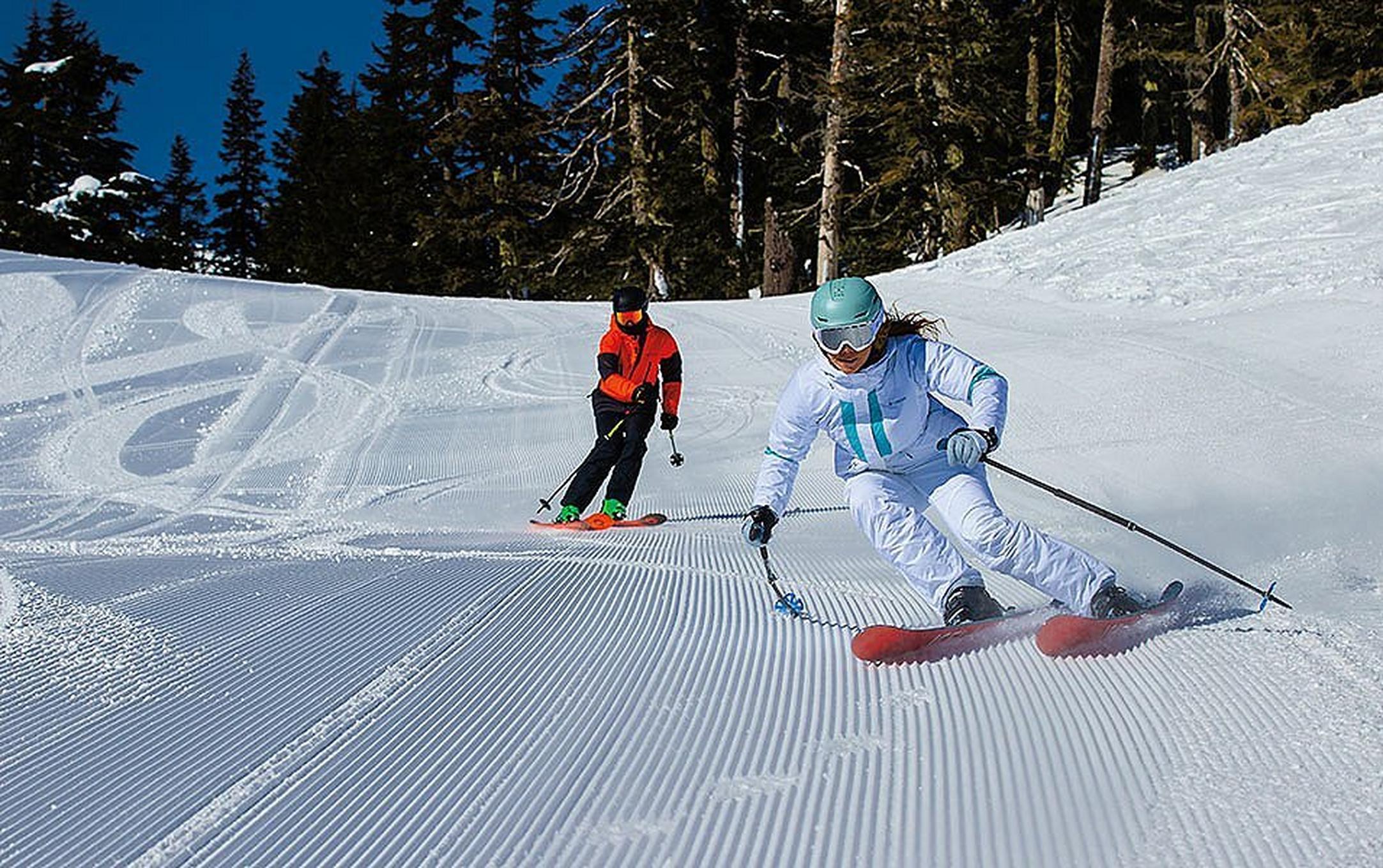 A woman in a white jacket with white pants skis down freshly groomed corduroy as a man in a red jacket and black pants skis behind her.