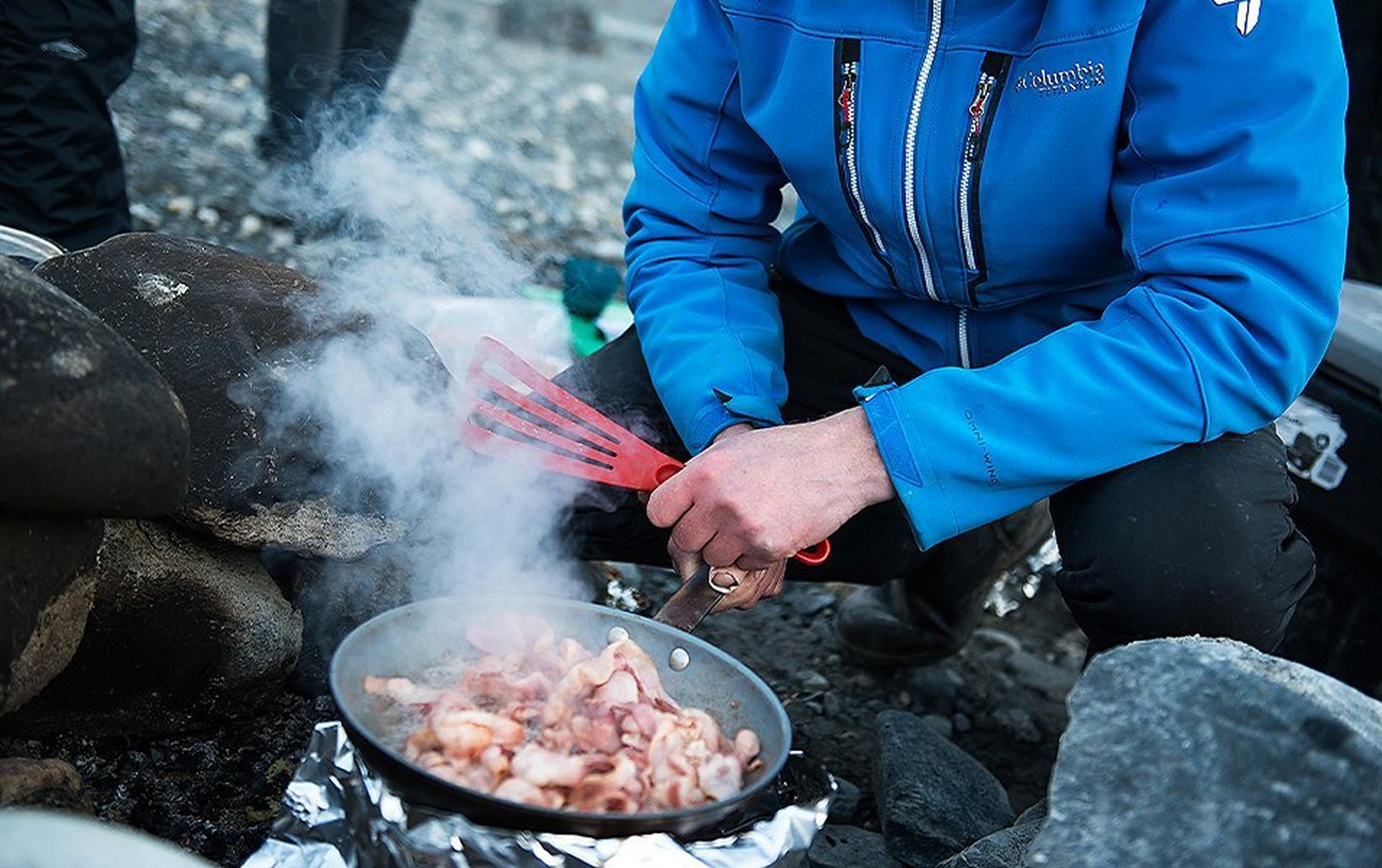 A winter camper fries bacon in a skillet over a fire.