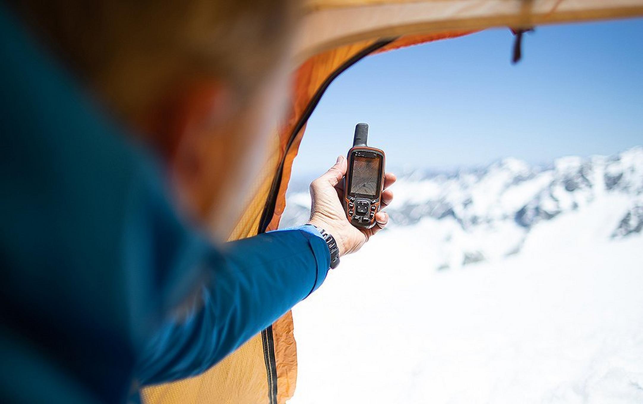 A man holds a GPS navigation device out of the window of a tent in a snowy setting.