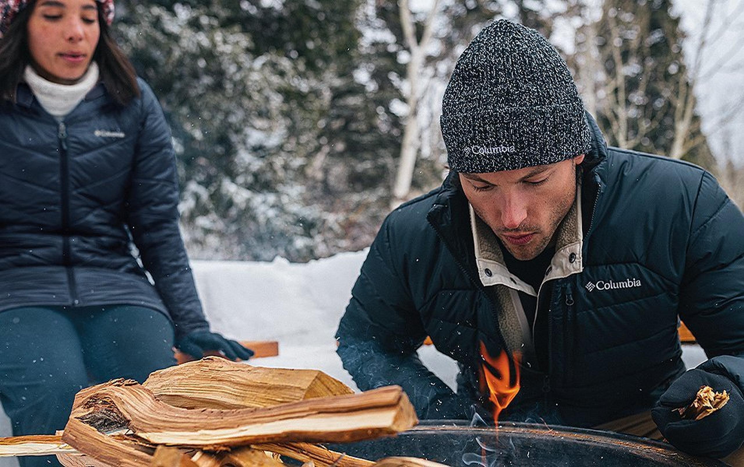 A man crouches down next to a campfire in a scenic winter setting as a woman looks on in the background.