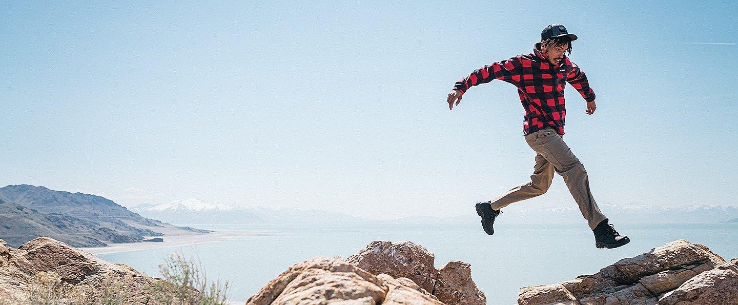 An African American man in a red-and-black checkered shirt and a black baseball cap jumps joyfully from rock to rock with a beautiful blue lake in the background.