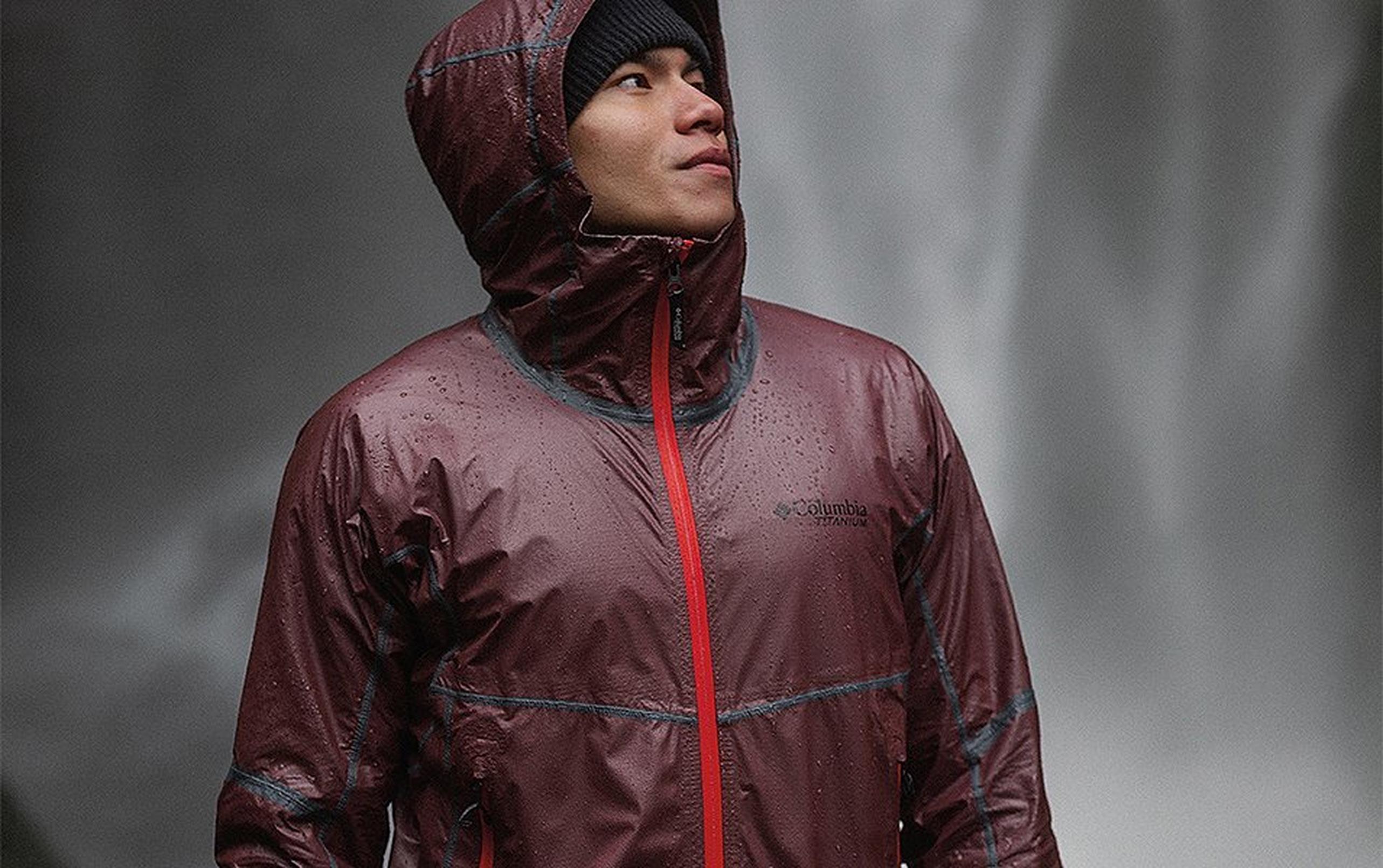 A man wearing a burgundy-colored Columbia Sportswear rain jacket stands in the rain in front of a cascading waterfall.