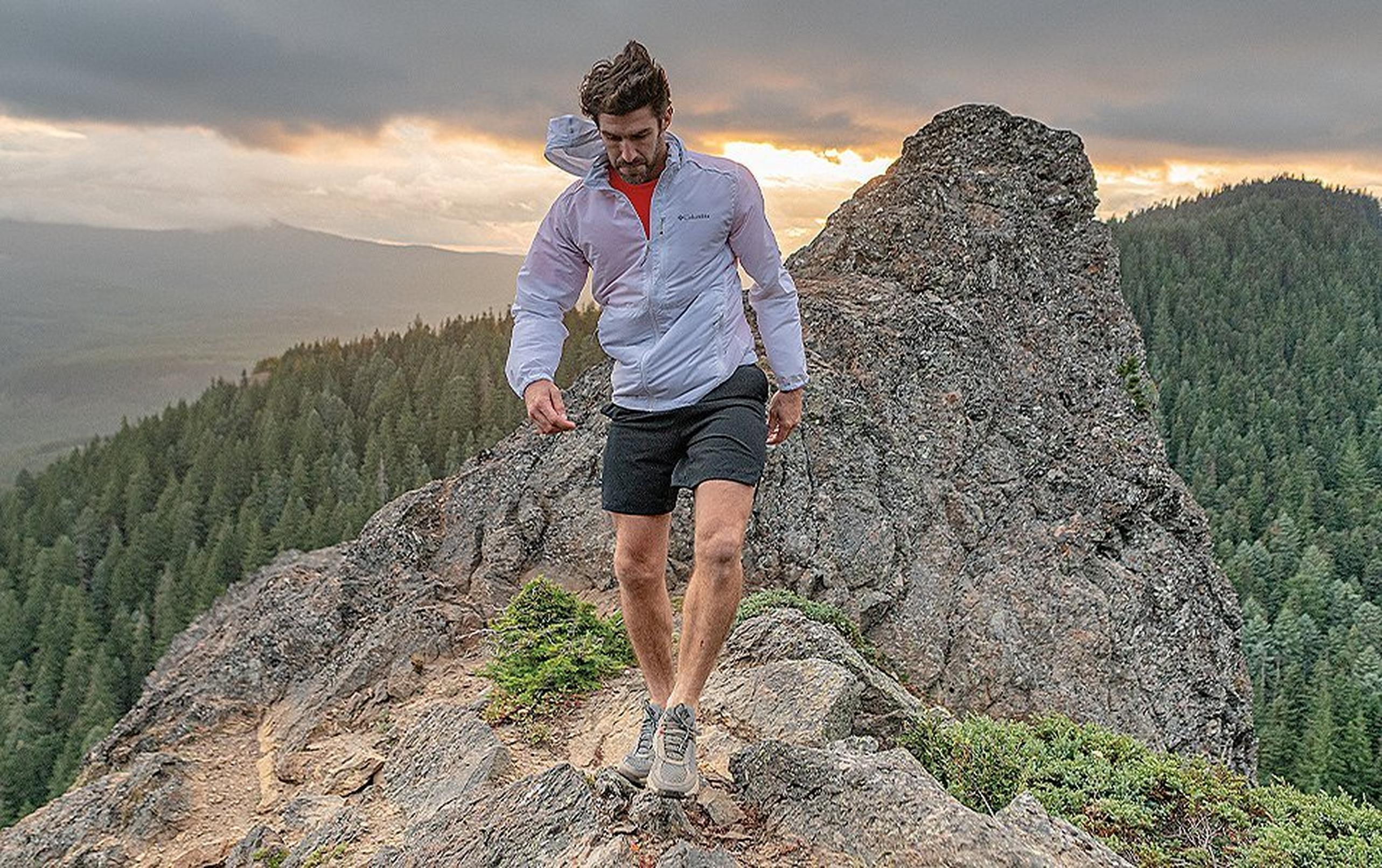 A man wearing a white Columbia Sportswear windbreaker walks along a rocky trail atop a beautiful mountain landscape.