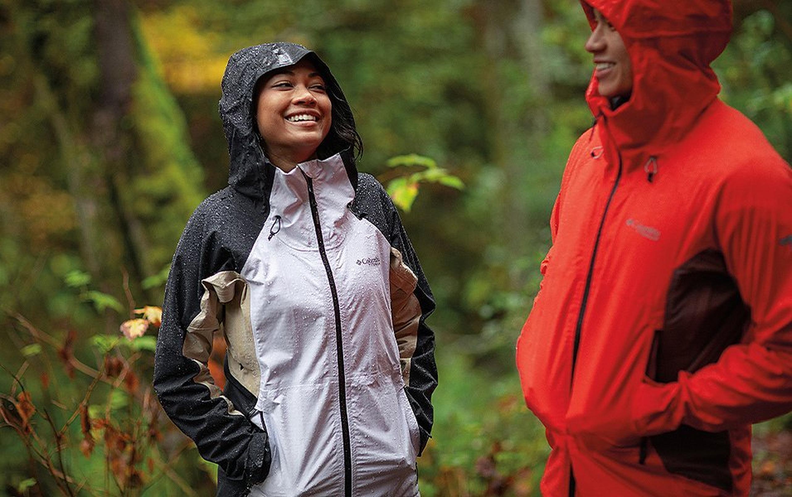 A woman stands in the rain in a forest smiling at a fellow hiker who is wearing a bright red rain coat.