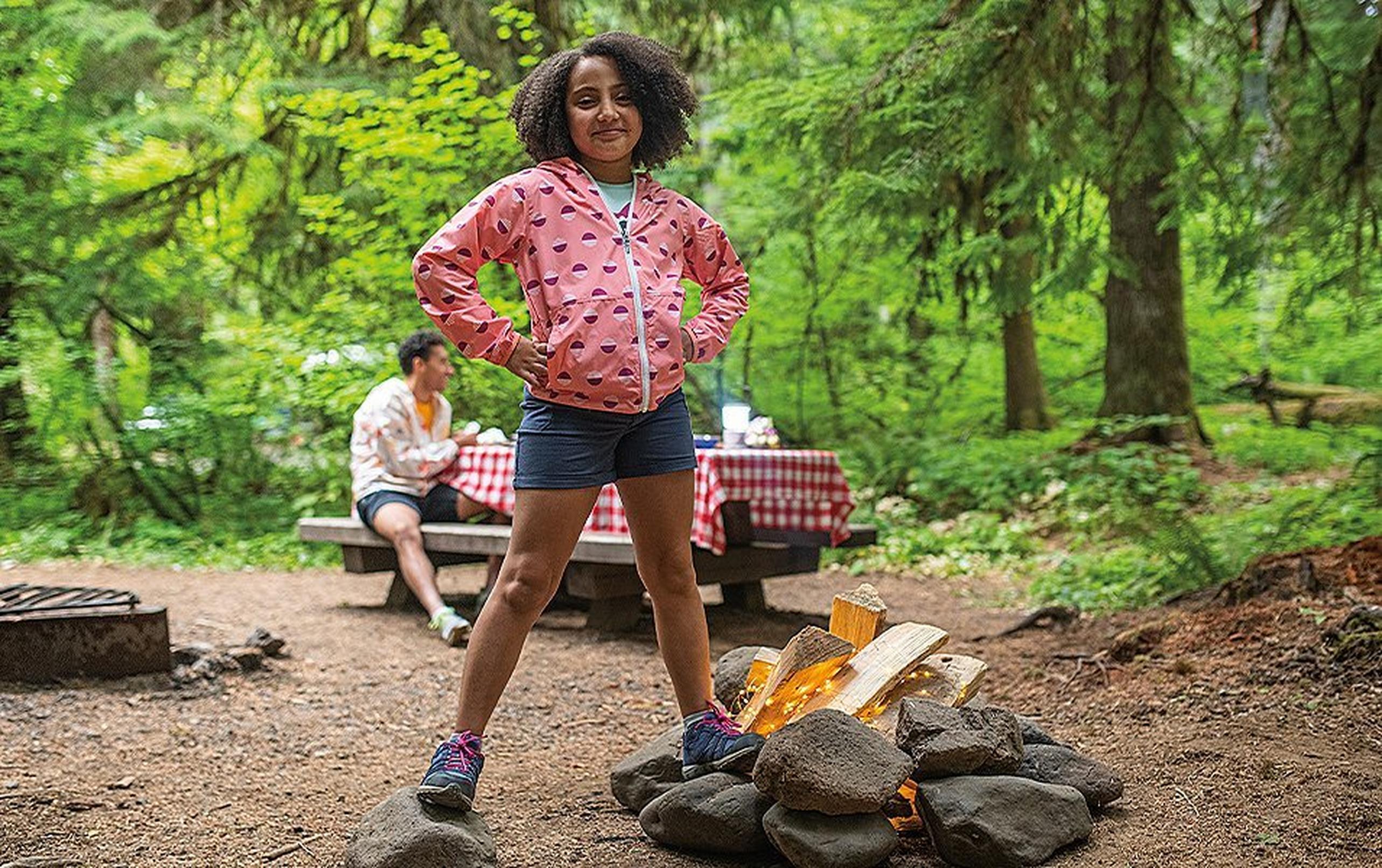 A girl wearing a pink windbreaker stands by a fire pit at a campground in a scenic forest setting.