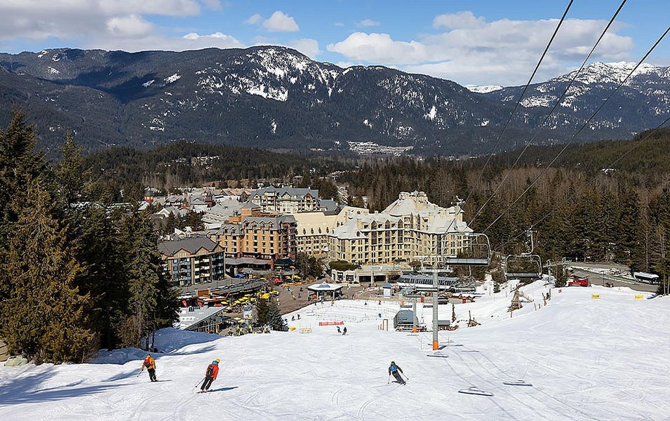 A wide shot of skiers heading down the slopes on a sunny day at Whistler Blackcomb with a ski lodge in the background.