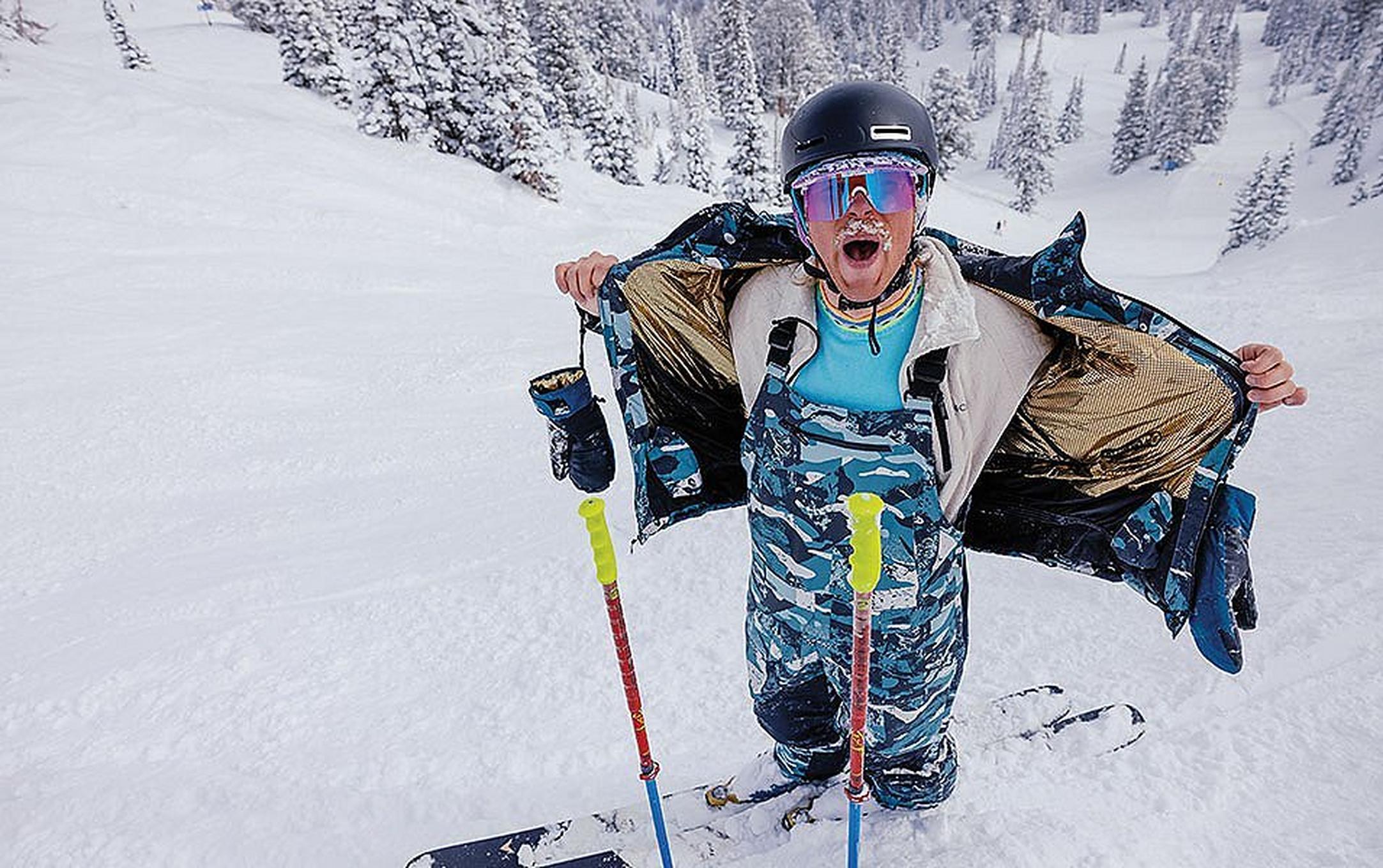 A skier wearing blue camo Columbia Sportswear bibs opens his jacket in a snowy powder field.