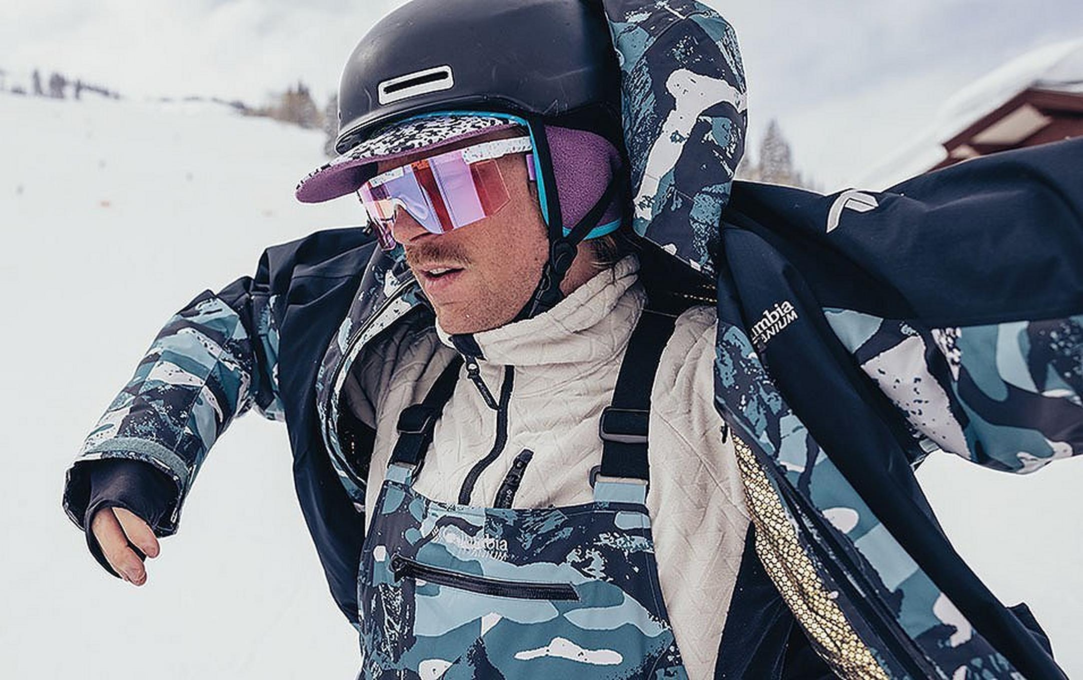 A skier wearing blue camo Columbia Sportswear bibs takes off his jacket in the middle of a ski slope.