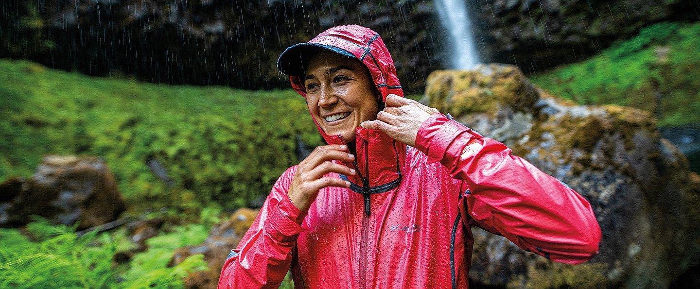 A woman wearing a bright red Columbia Sportswear rain jacket smiles widely as she reaches for her collar with green foliage and rocks in the background.