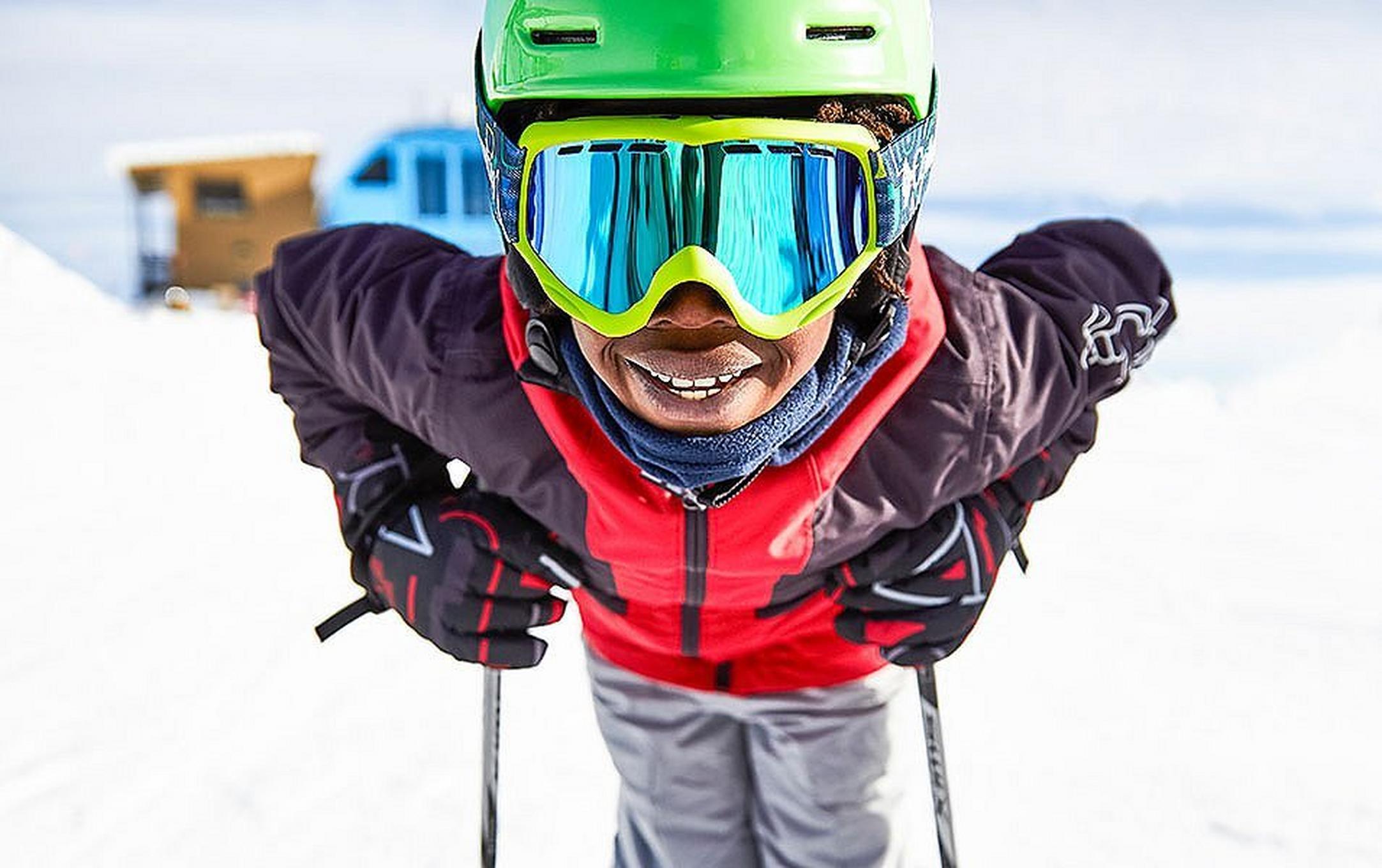 A young skier standing in the snow leans forward and smiles big for the camera on a gorgeous sunny day on the mountain.