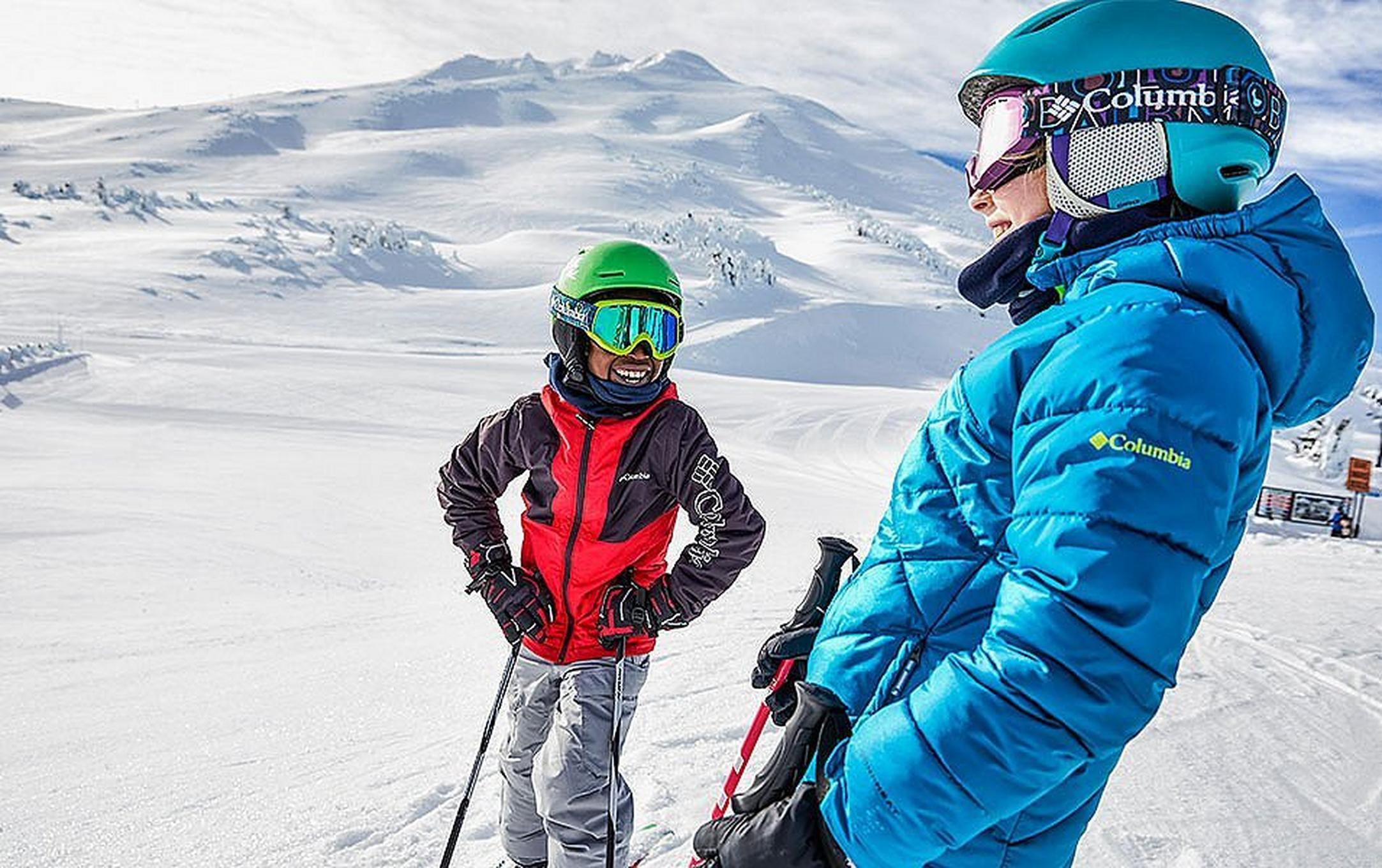 A skier wearing a blue Columbia Sportswear jacket smiles as he talks to a youth standing next to him on a sunny day on a snow-covered mountain.
