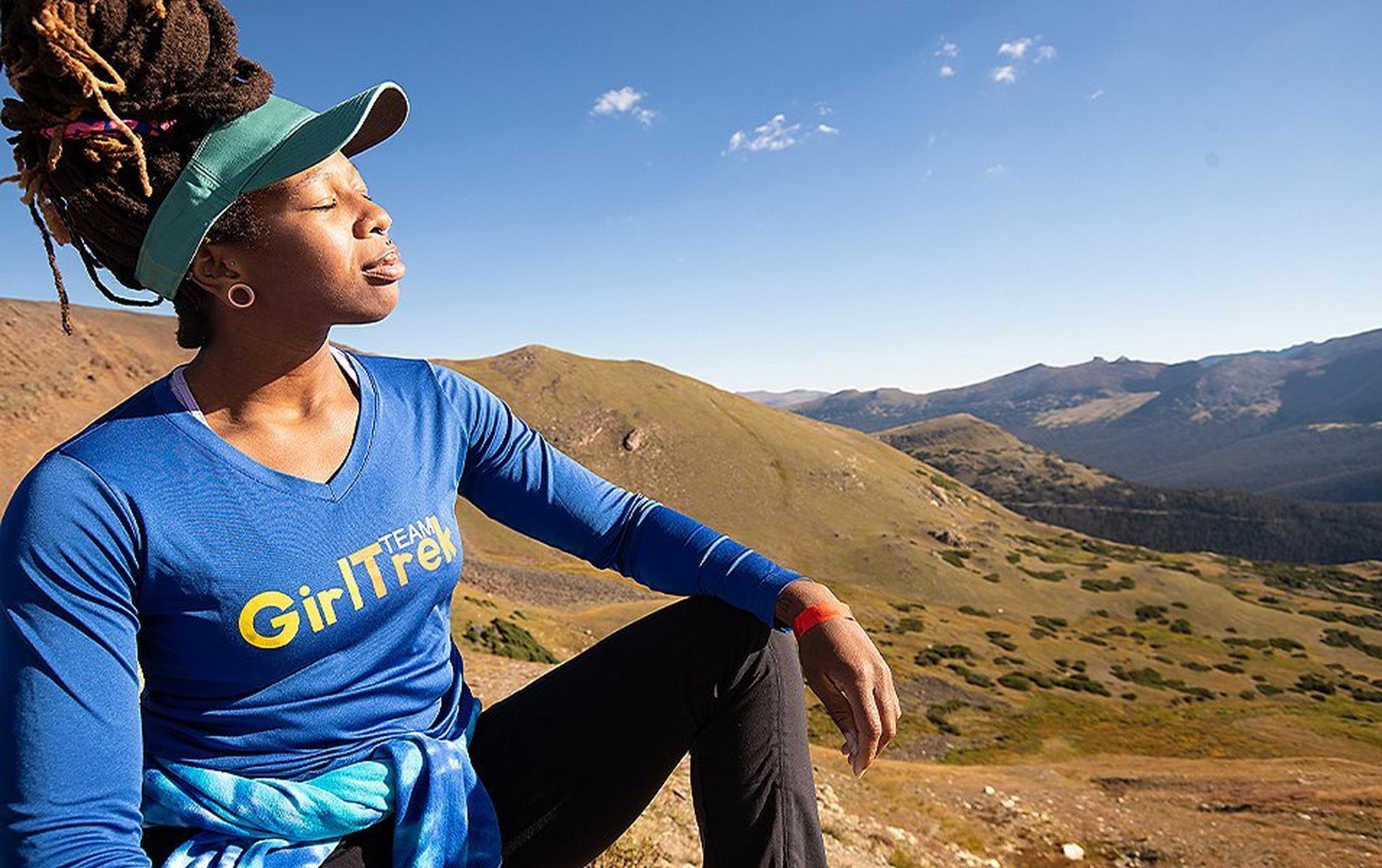 An African American woman wearing a blue GirlTrek jacket basks in the sun in the middle of a beautiful landscape.