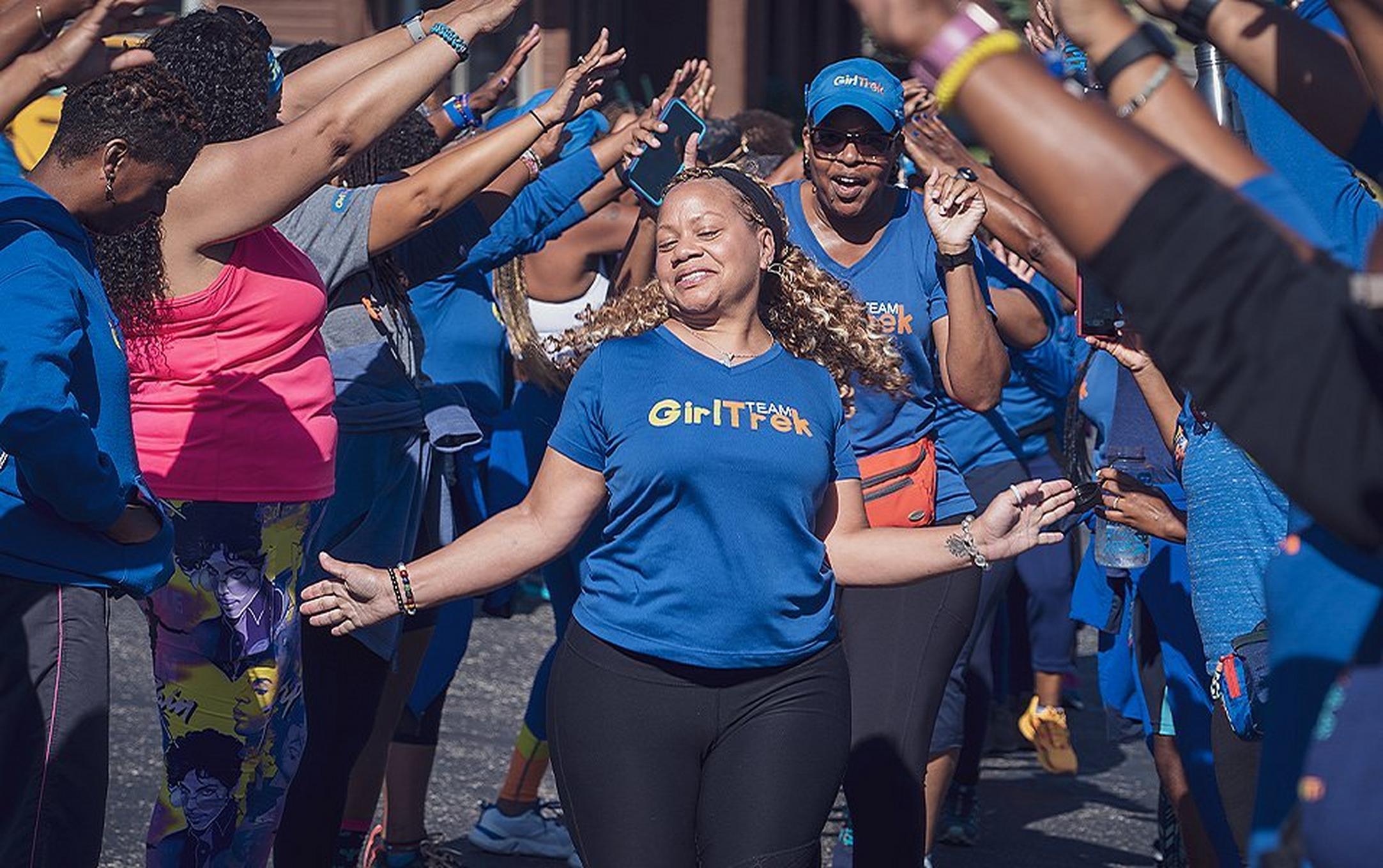 A woman in a blue GirlTrek shirt walks towards the camera enthusiastically as a group of women stand around her making a bridge.
