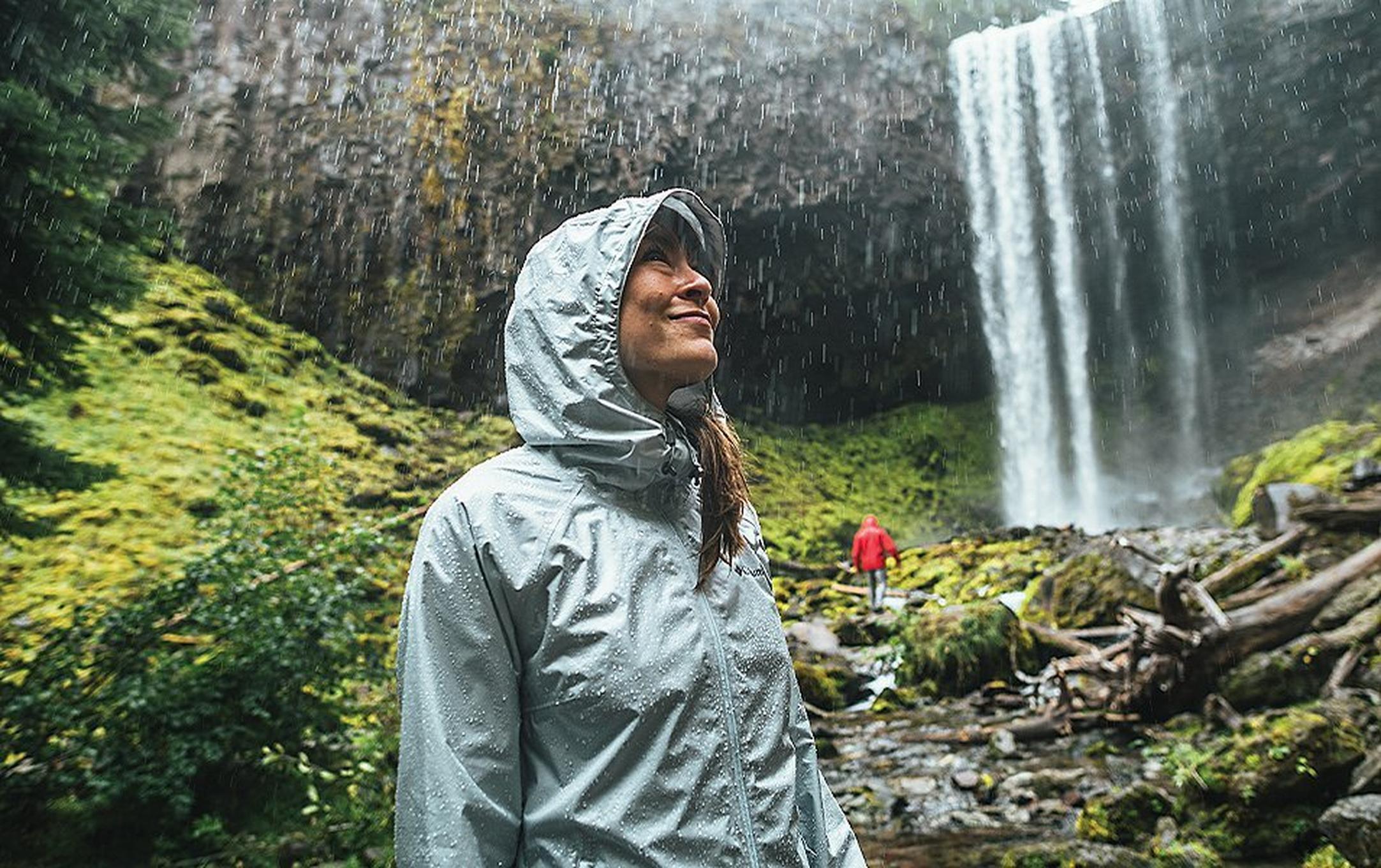 Close up of woman standing in the rain near a waterfall with a man farther in the background.