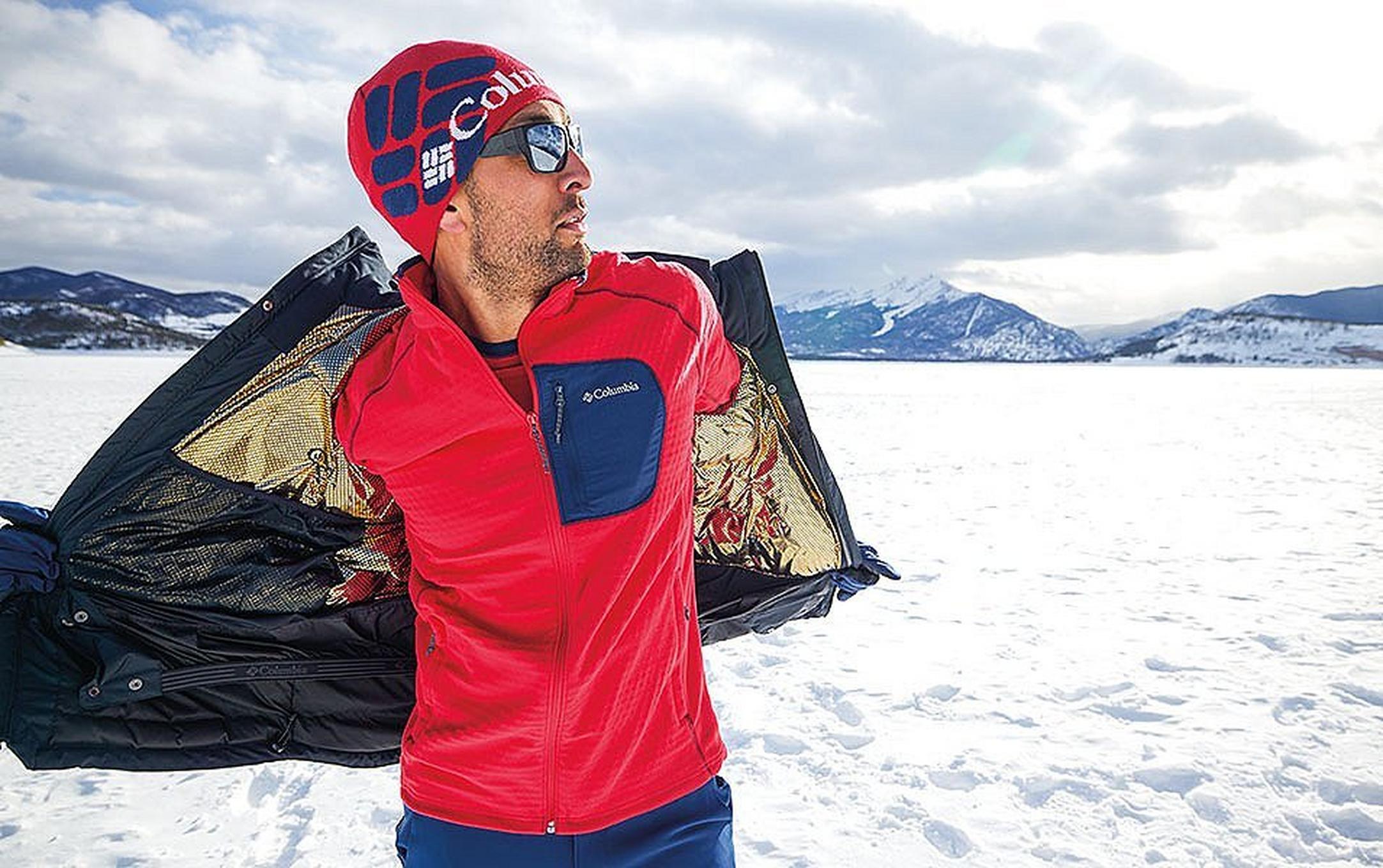 A man stands in a scenic snowy setting taking off a Columbia Sportswear jacket with a layer of red fleece underneath.