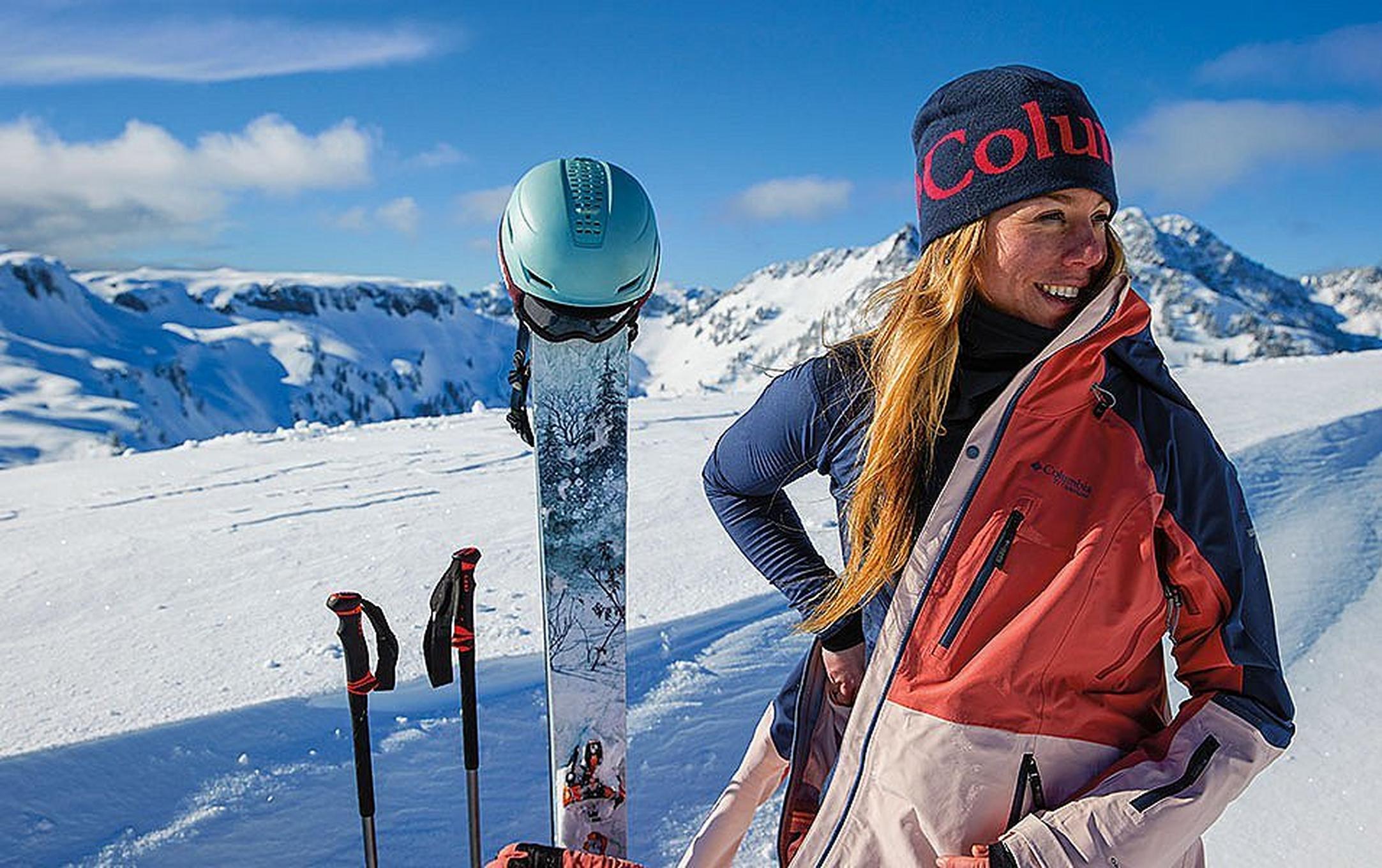 A skier stands on a snow-covered mountain taking off her ski jacket as her skis, poles, helmet, and backpack stick out of the snow next to her. Underneath her ski coat she is wearing a warm Columbia Sportswear baselayer.