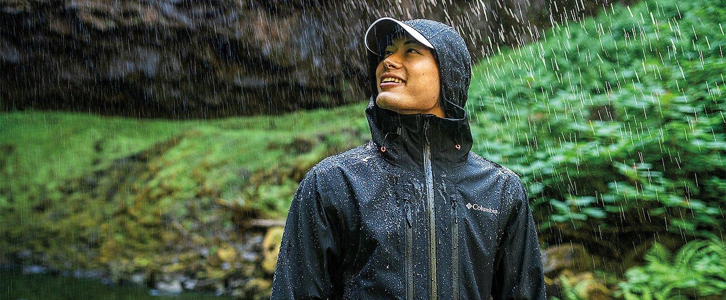 A man in a black Columbia Sportswear jacket leans to the side smiling in the rain with lush green foliage in the background.
