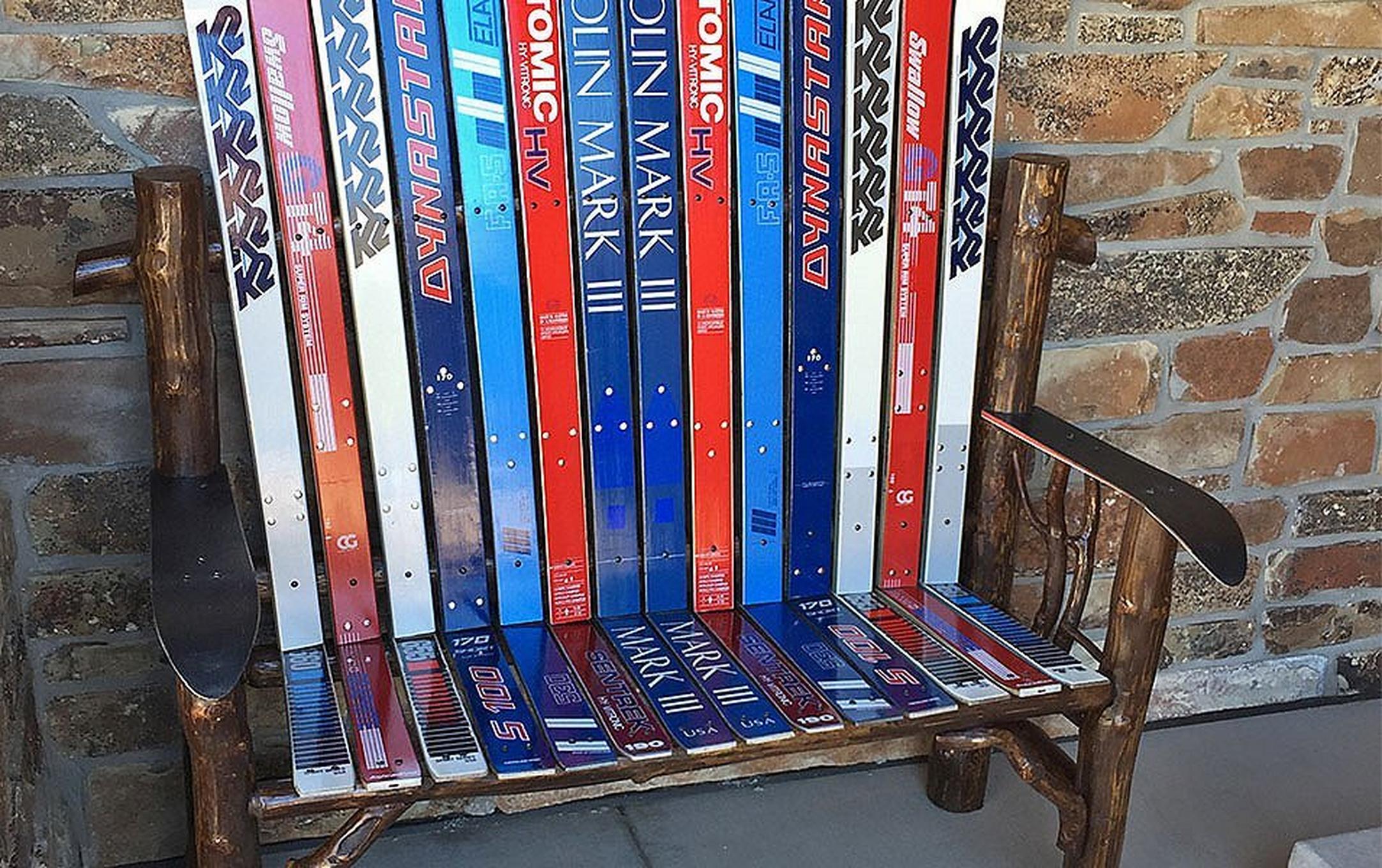 A patio bench made from colorful skis sits outside in a concrete area in front of a brick wall.