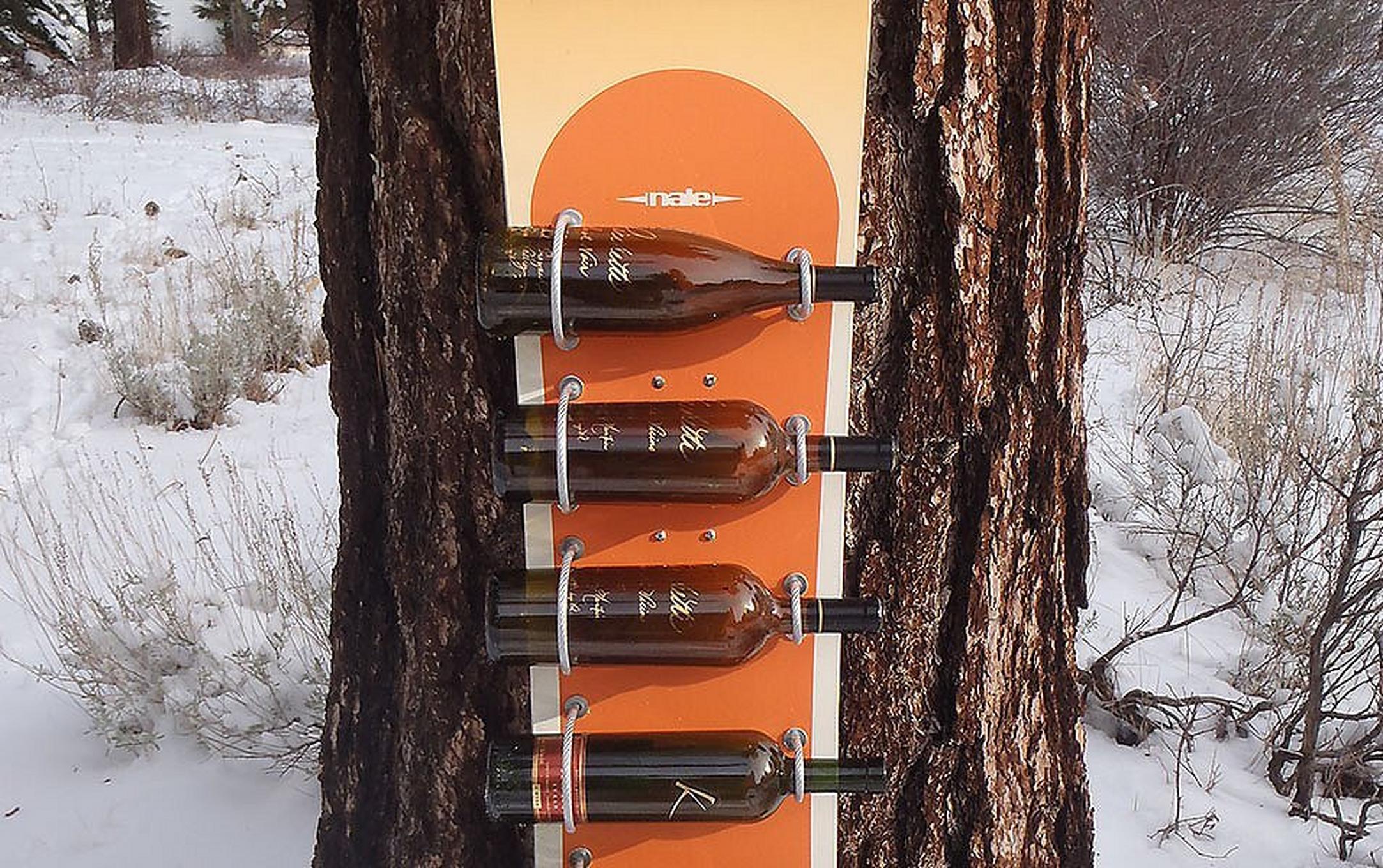 A wall-mounted wine rack made from an upcycled snowboard leans against a tree in a snowy outdoor setting.
