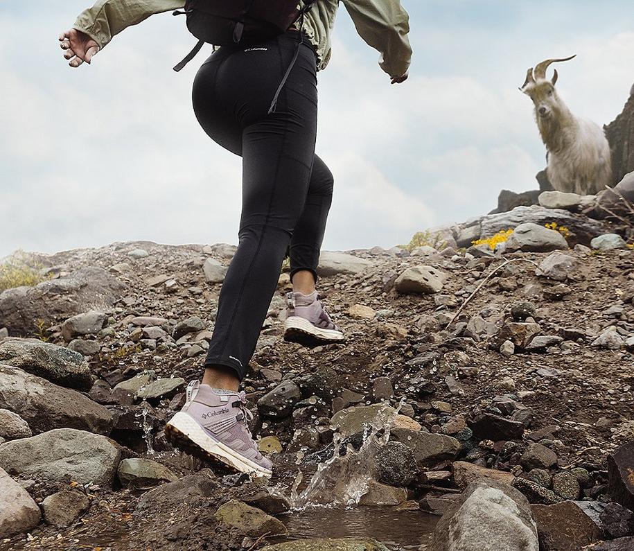 A man hikes up a rocky trail wearing Columbia Sportswear’s Facet 75 Mid shoe as a mountain goat watches in the distance.