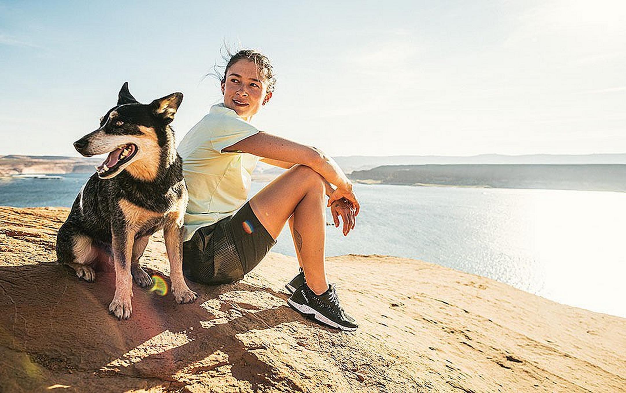 A woman sits on a rocky overlook above a sunny blue body of water with a smiling black dog at her side.