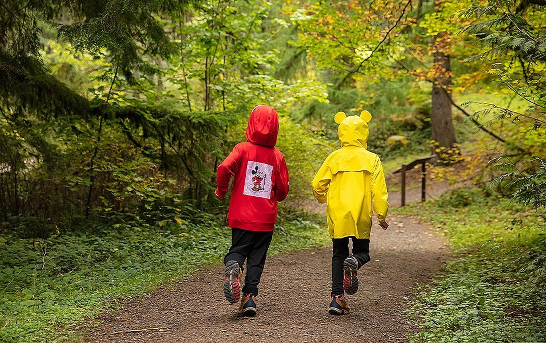 Two kids in rain coats running on a dirt path.
