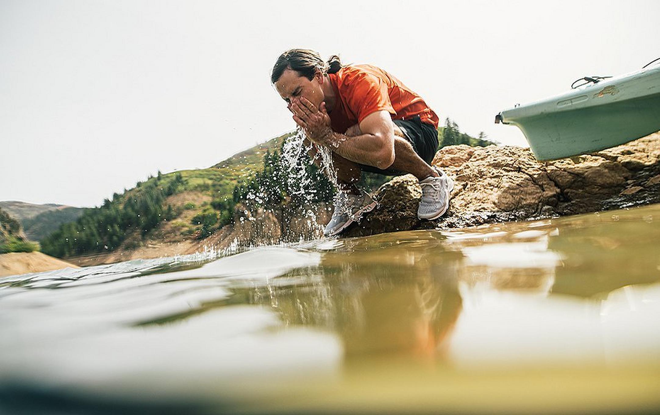 A man in a red shirt squats next to a river splashing water into his mouth.