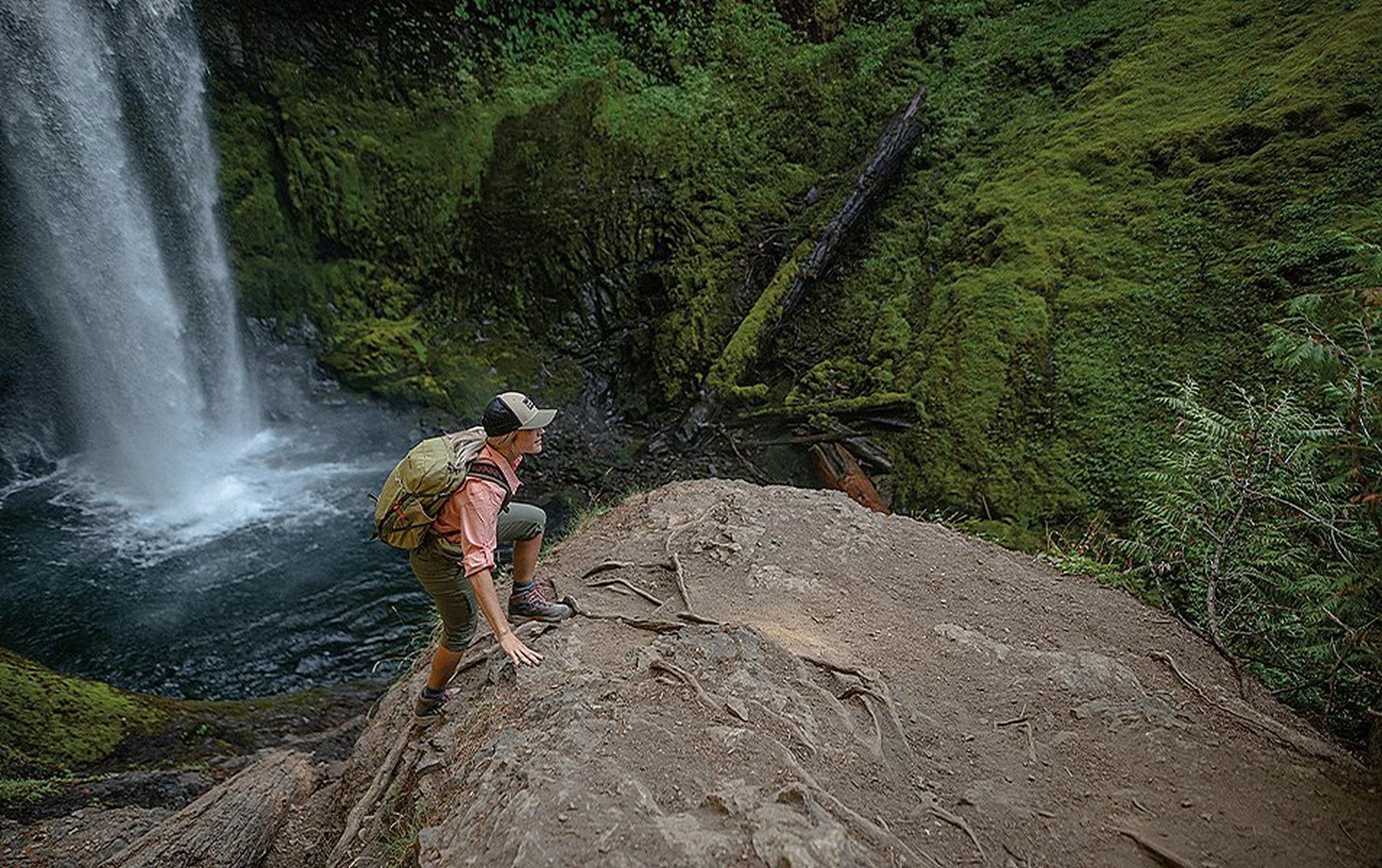 A woman in a pink shirt with a baseball cap climbs up a muddy trail in the woods with a waterfall in the background.