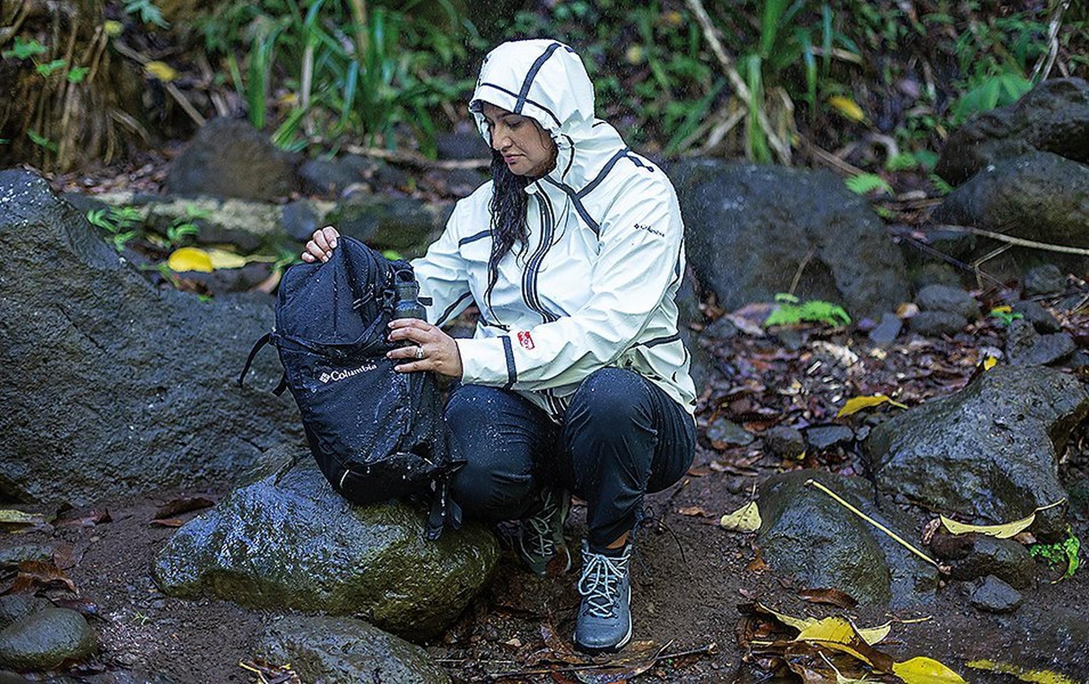 Woman holding a backpack sitting on a rock in the woods.
