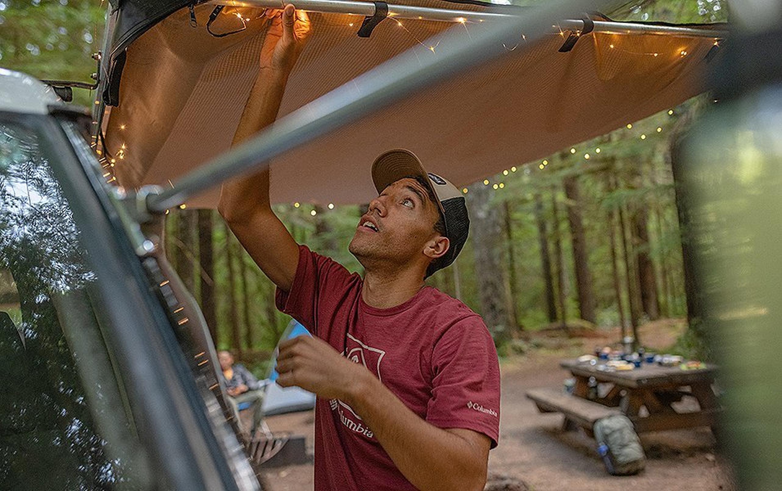 A man adjusts camping lighting on the back of his vehicle in a scenic forest campsite.