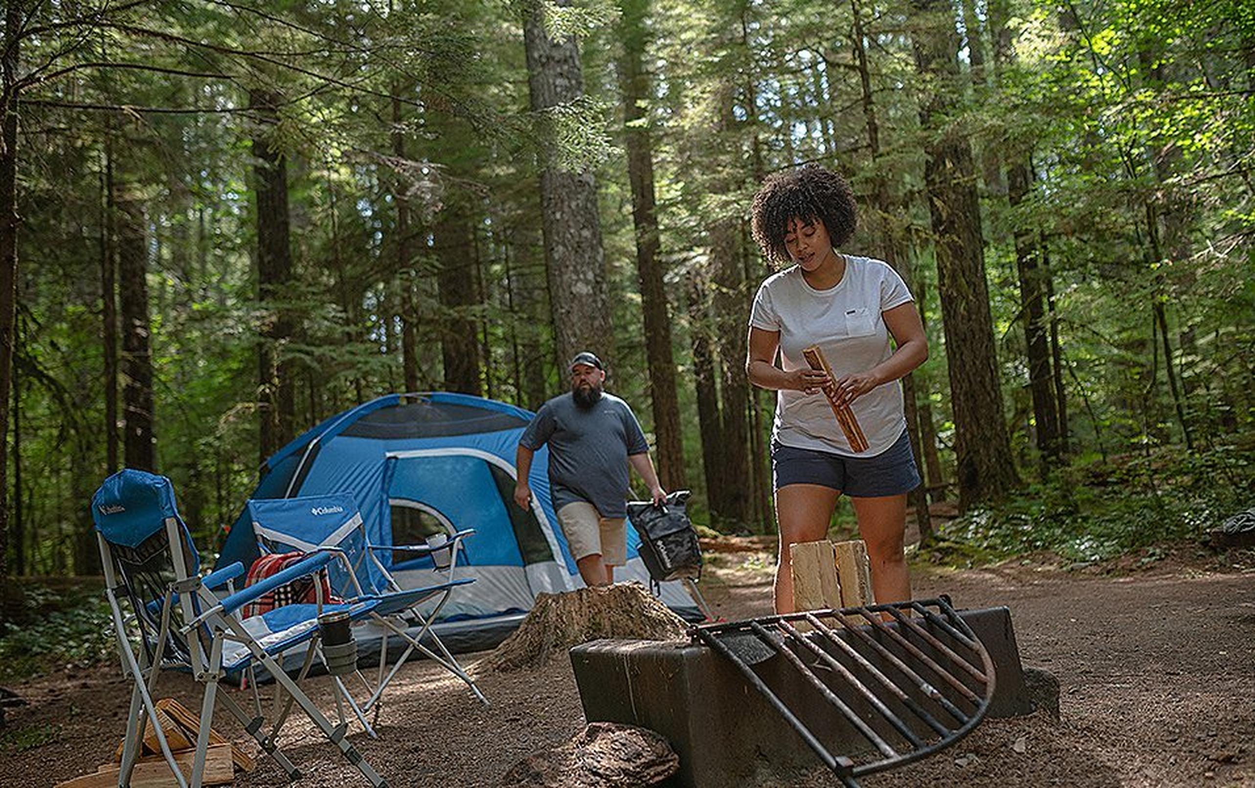 A woman gets a campfire started in the forest as a man walks in front of the tent behind her.