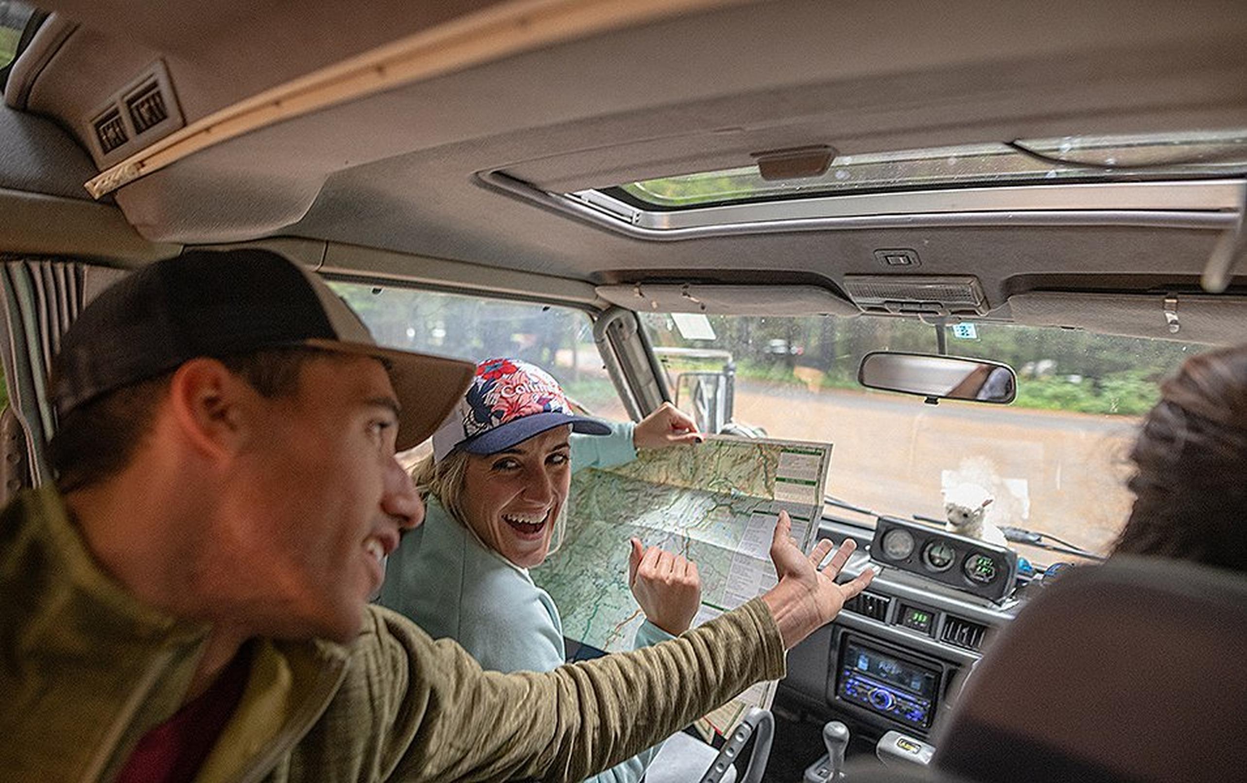 A woman in a car laughs in the driver’s seat as she reads a map while a man in the back seat reaches an arm forward.