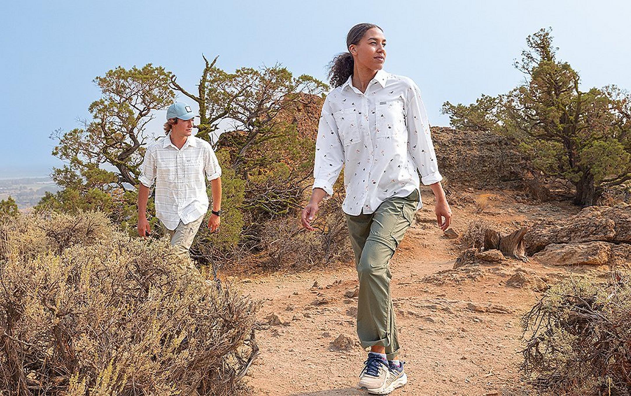 A woman wearing a UPF-rated Columbia Sportswear Silver Ridge shirt hikes through a high desert landscape with a man in UPF clothes trekking behind her.
