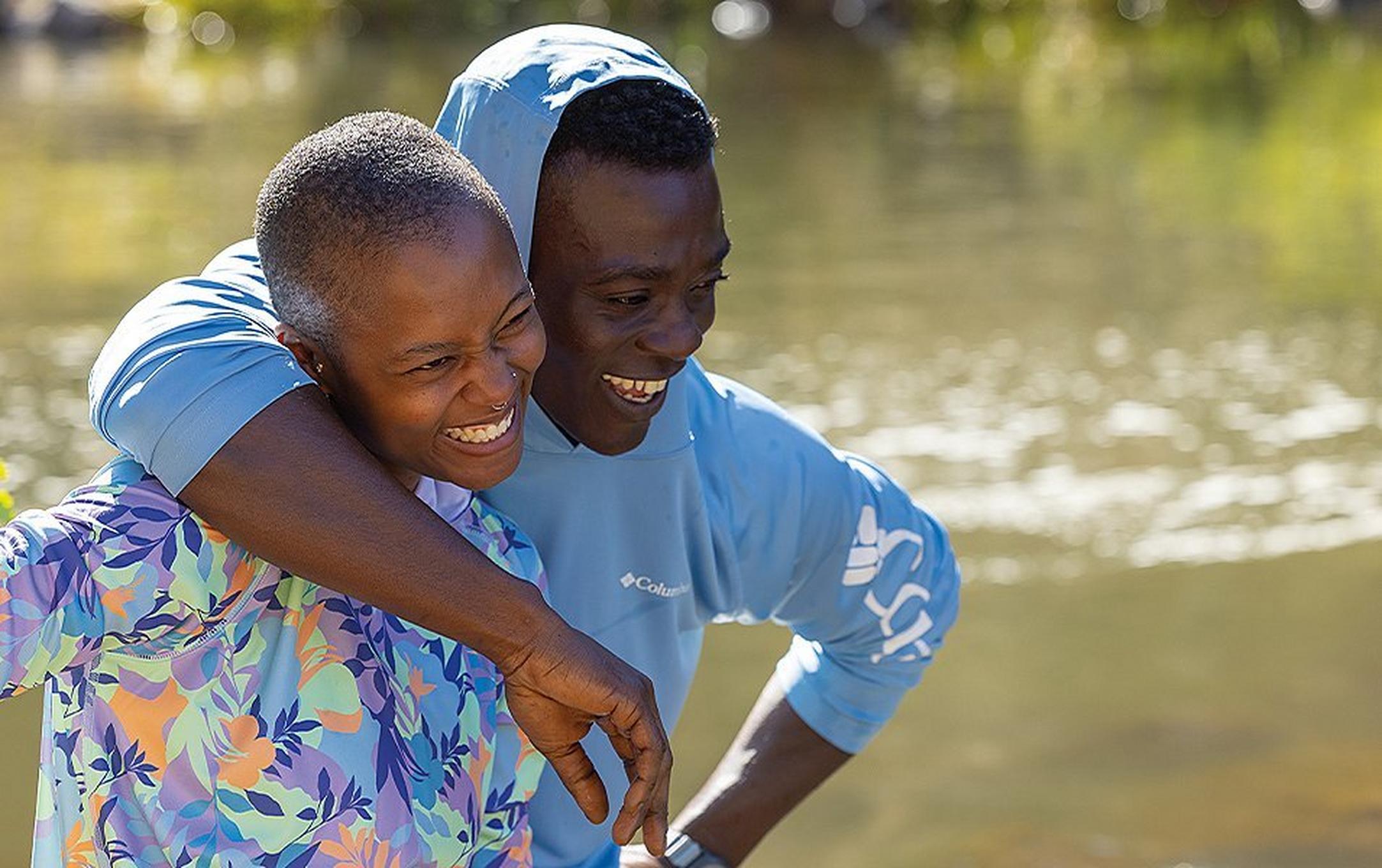 A man wearing an UPF shirt puts his arm around a kid and smiles with a scenic river in the background.