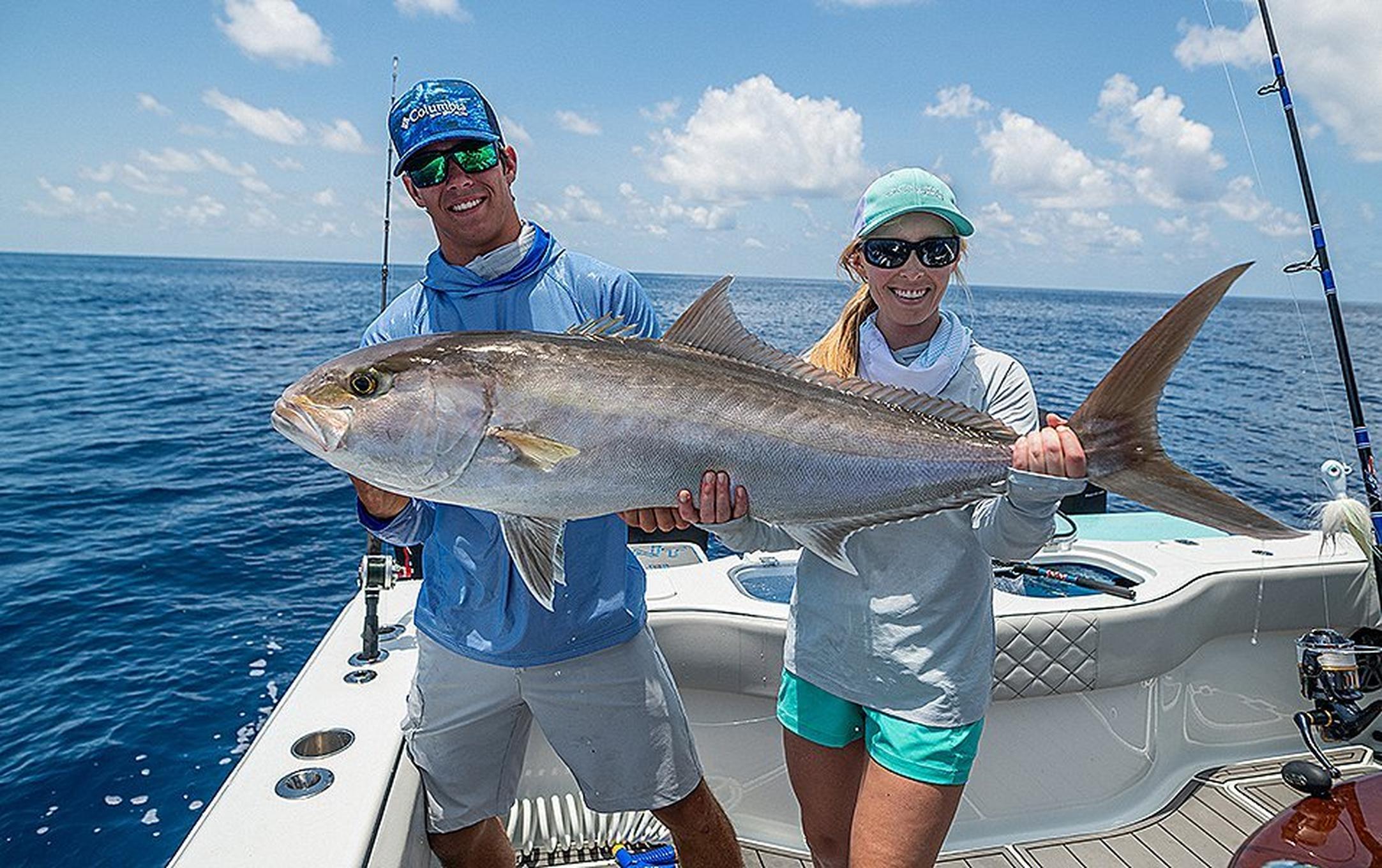 A young man in a blue long-sleeved Columbia Sportswear Terminal Tackle shirt holds up a giant fish on a boat next to a young woman wearing a similar long-sleeved white shirt. They are both wearing baseball caps and smiling.