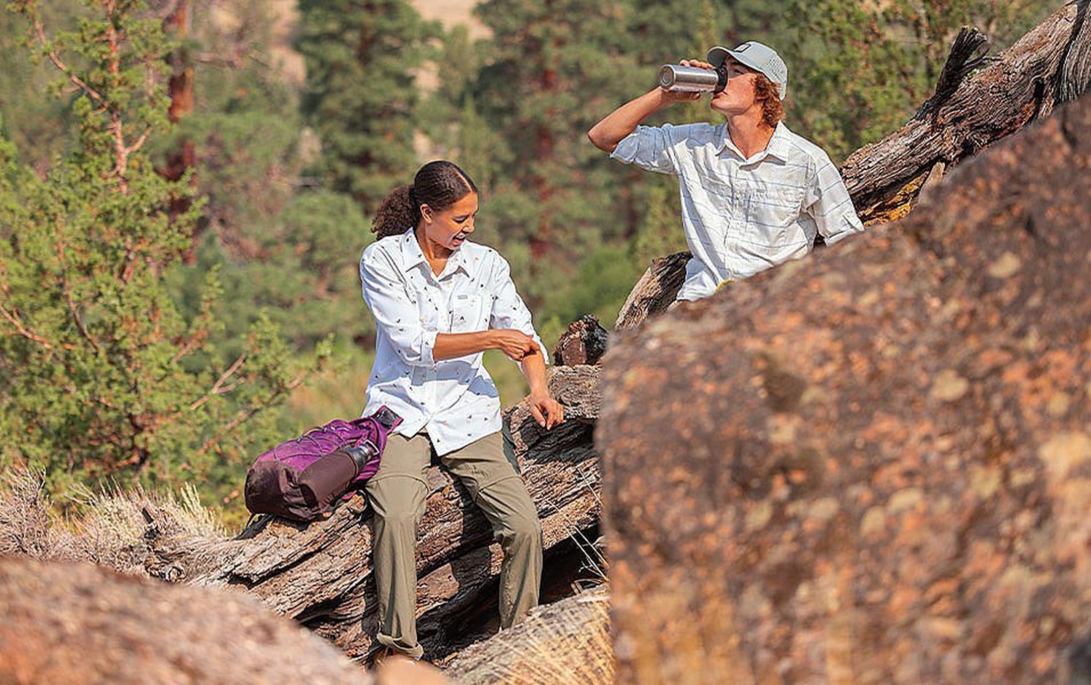 A woman rolls up her sleeve while a young man next to her takes a sip from his water bottle as the two hikers pause on the trail.
