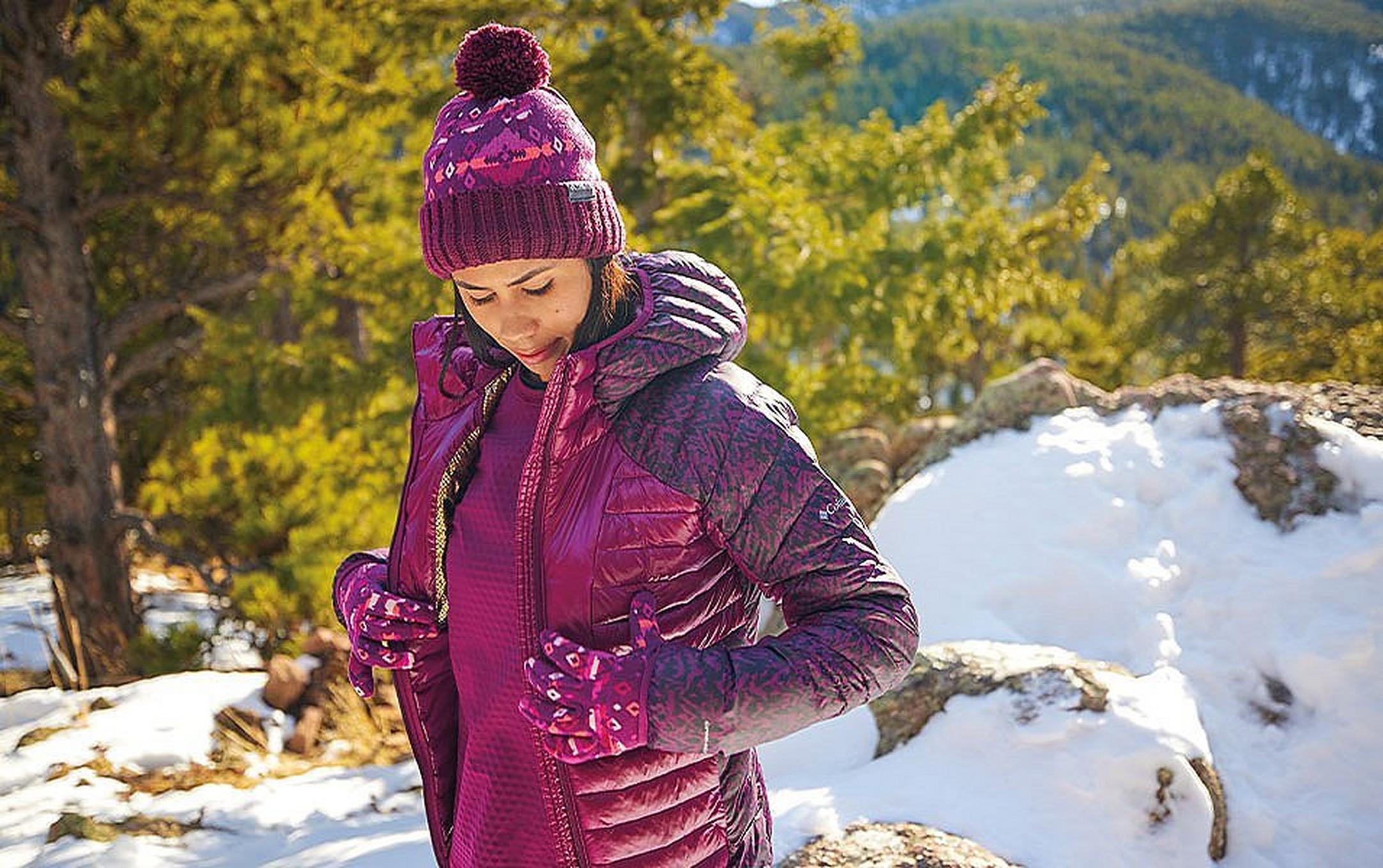 A woman wearing a fleece and puffer jacket stands on a scenic winter hiking trail looking down at the ground.