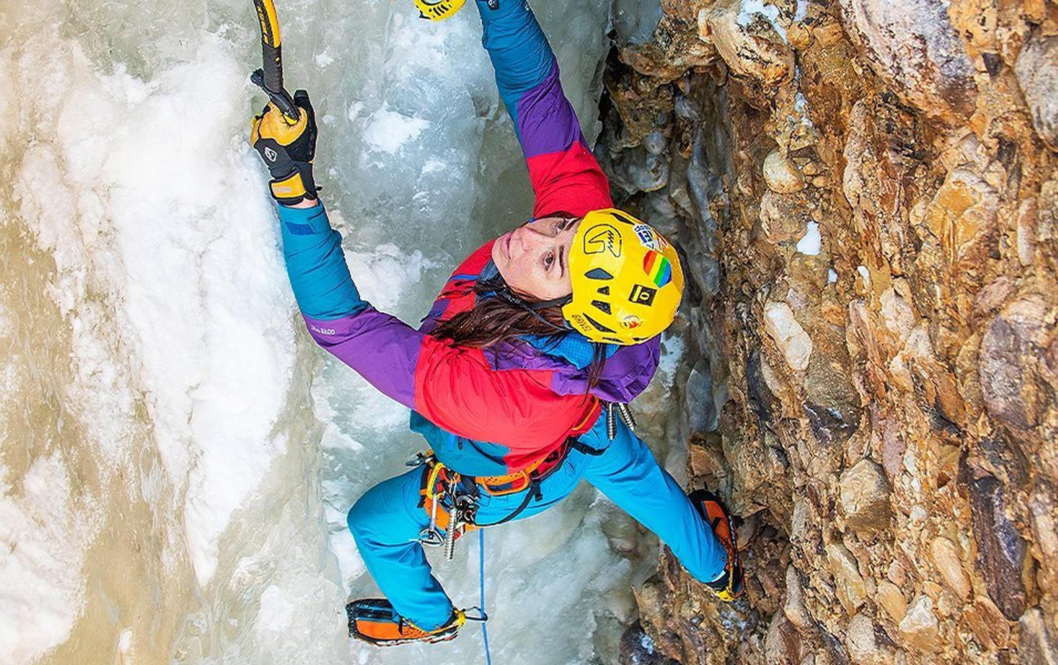 Pro climber Nikki Smith scales a vertical rock face covered in ice while wearing a bright multi-colored jacket and yellow helmet.