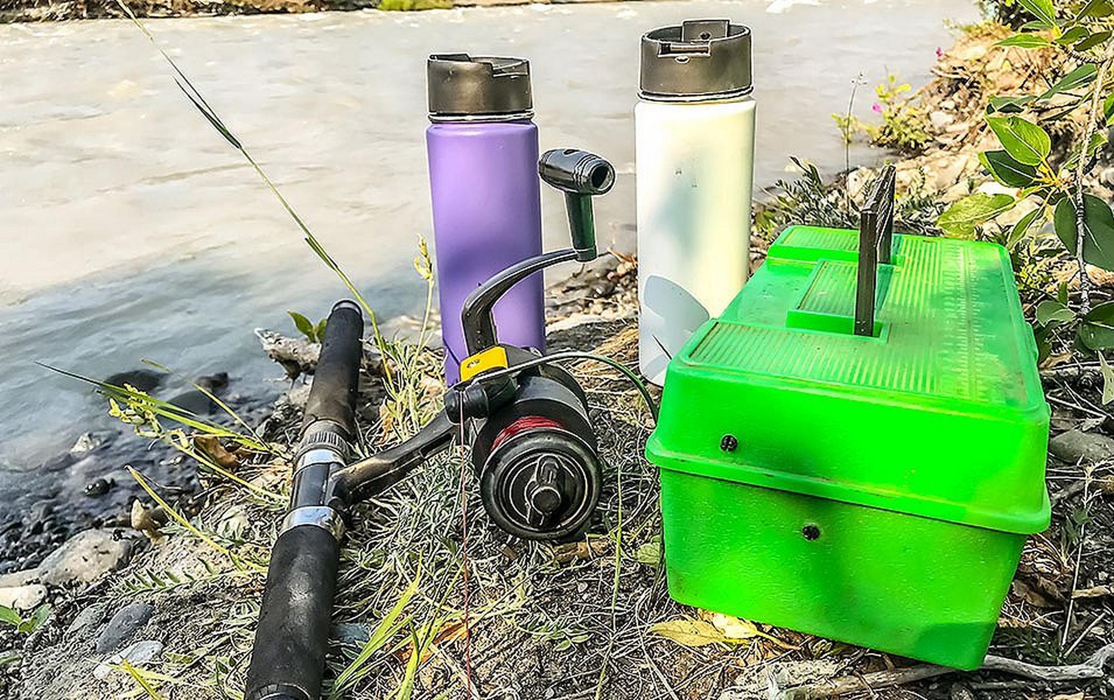 A close-up shot of a pile of fishing gear sitting by a river bank that includes a fishing rod, water bottles, and tackle box.