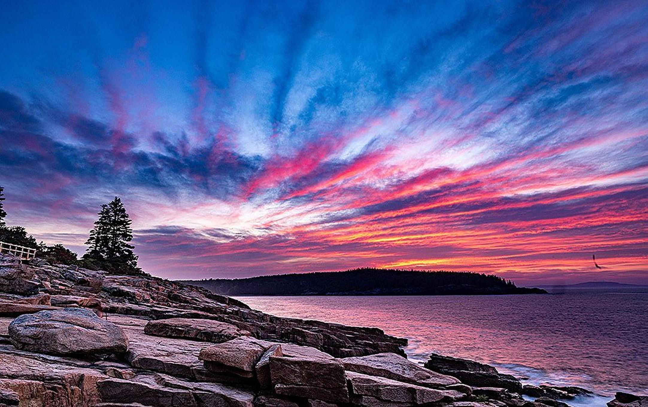 A dramatic blue, purple and rose sunset illuminates big flat rocks extending to the shoreline in Acadia National Park.