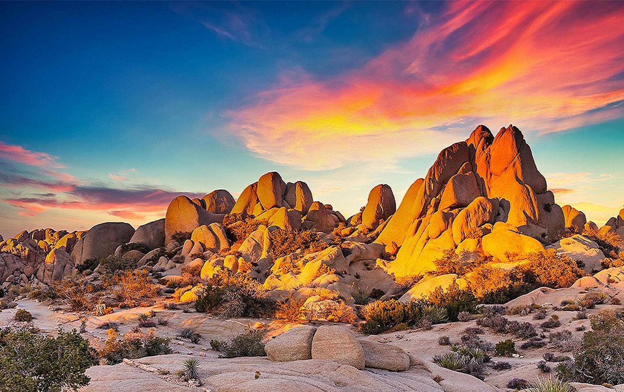 Giant boulders dot the desert landscape in Joshua Tree National Park as the sun sets in the background.