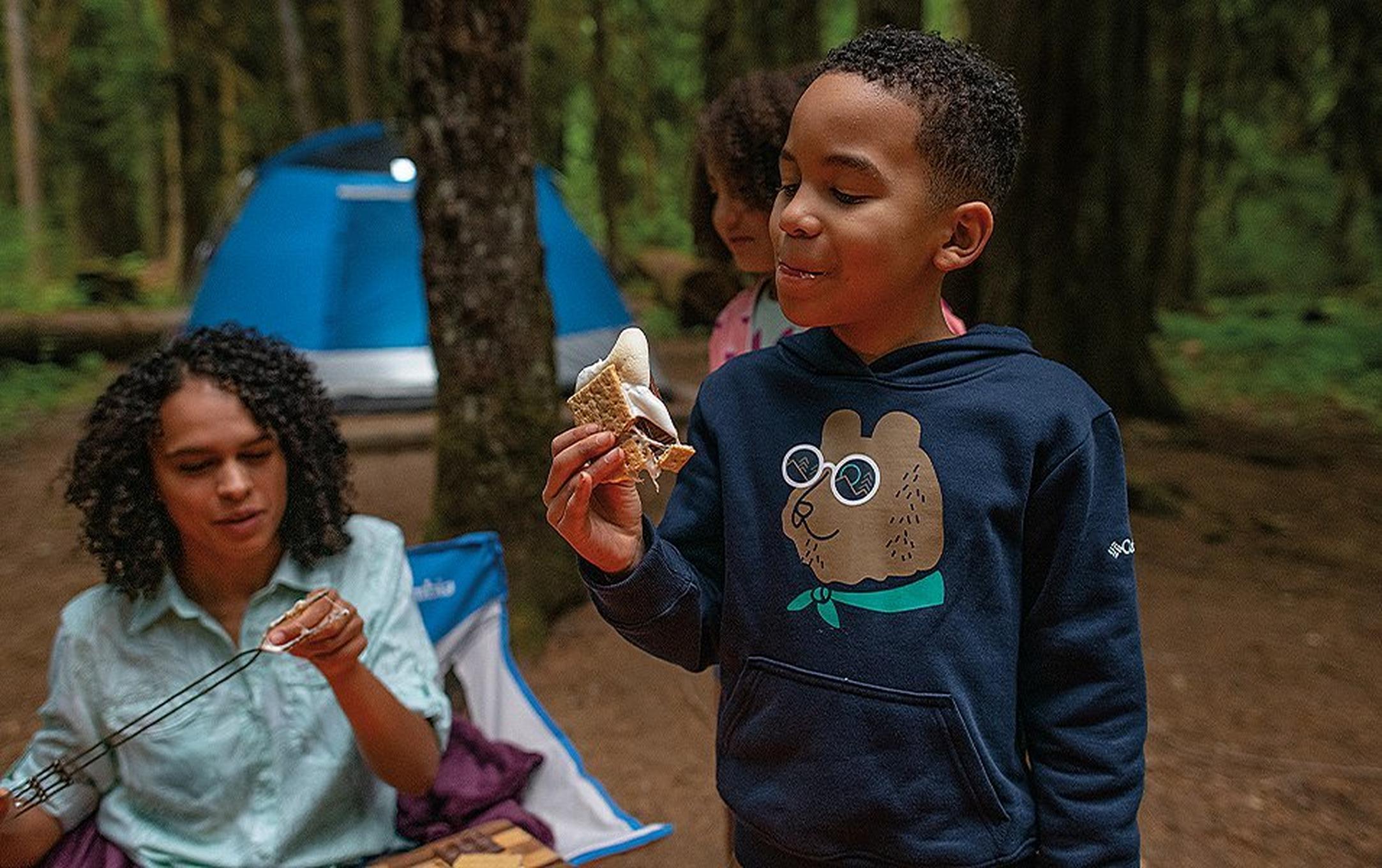 A boy in a navy blue sweatshirt eats S’mores in the woods as his mom skewers a marshmallow in the background.