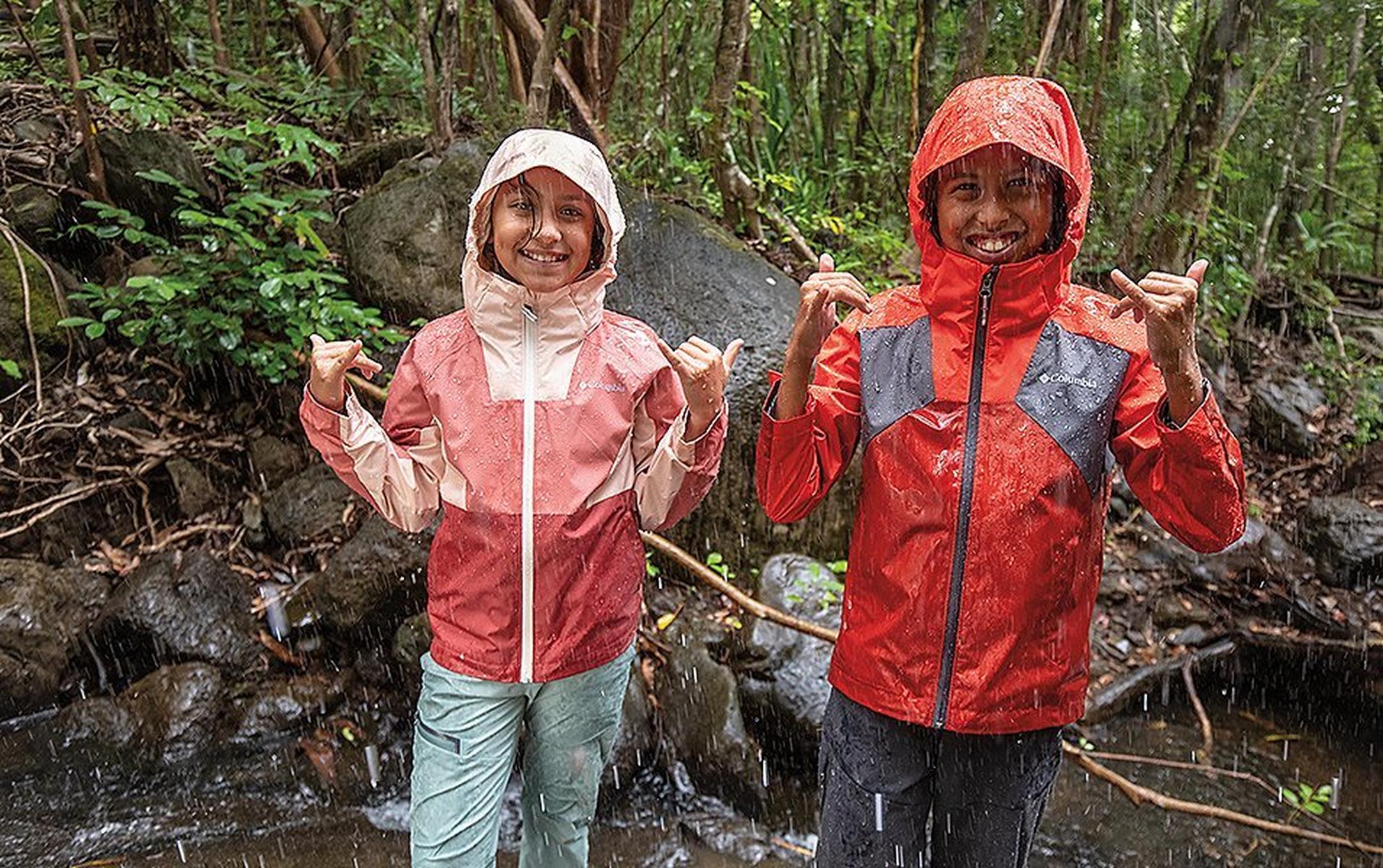Two girls wearing Columbia Sportswear jackets stand in the rain smiling in the middle of the forest.