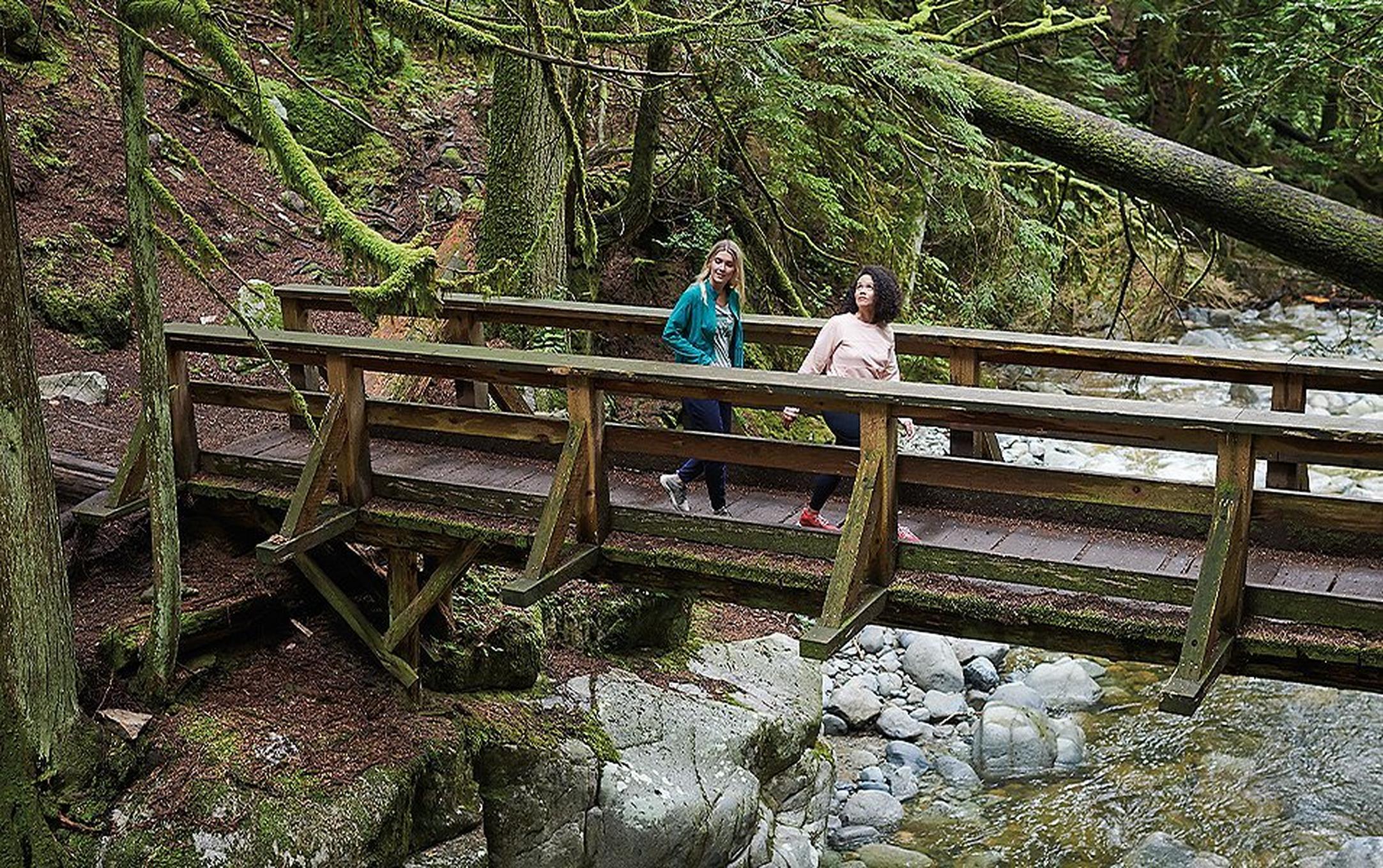 Two women hike across a wooden bridge in a mossy forest with a rushing creek beneath them.