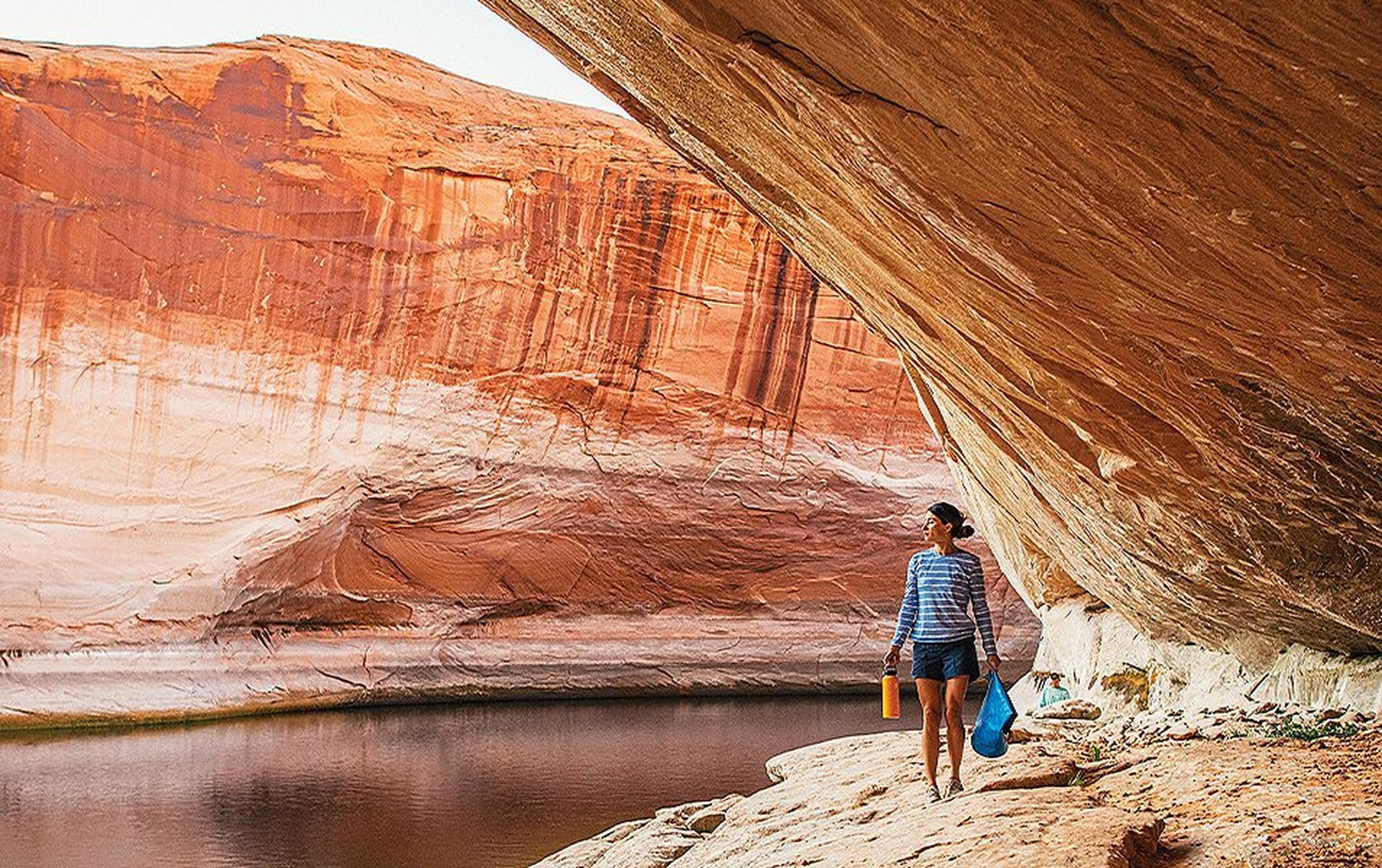 A woman in a blue striped shirt hikes through a stunning slot canyon with a yellow water bottle in one hand and a blue dry bag in the other.