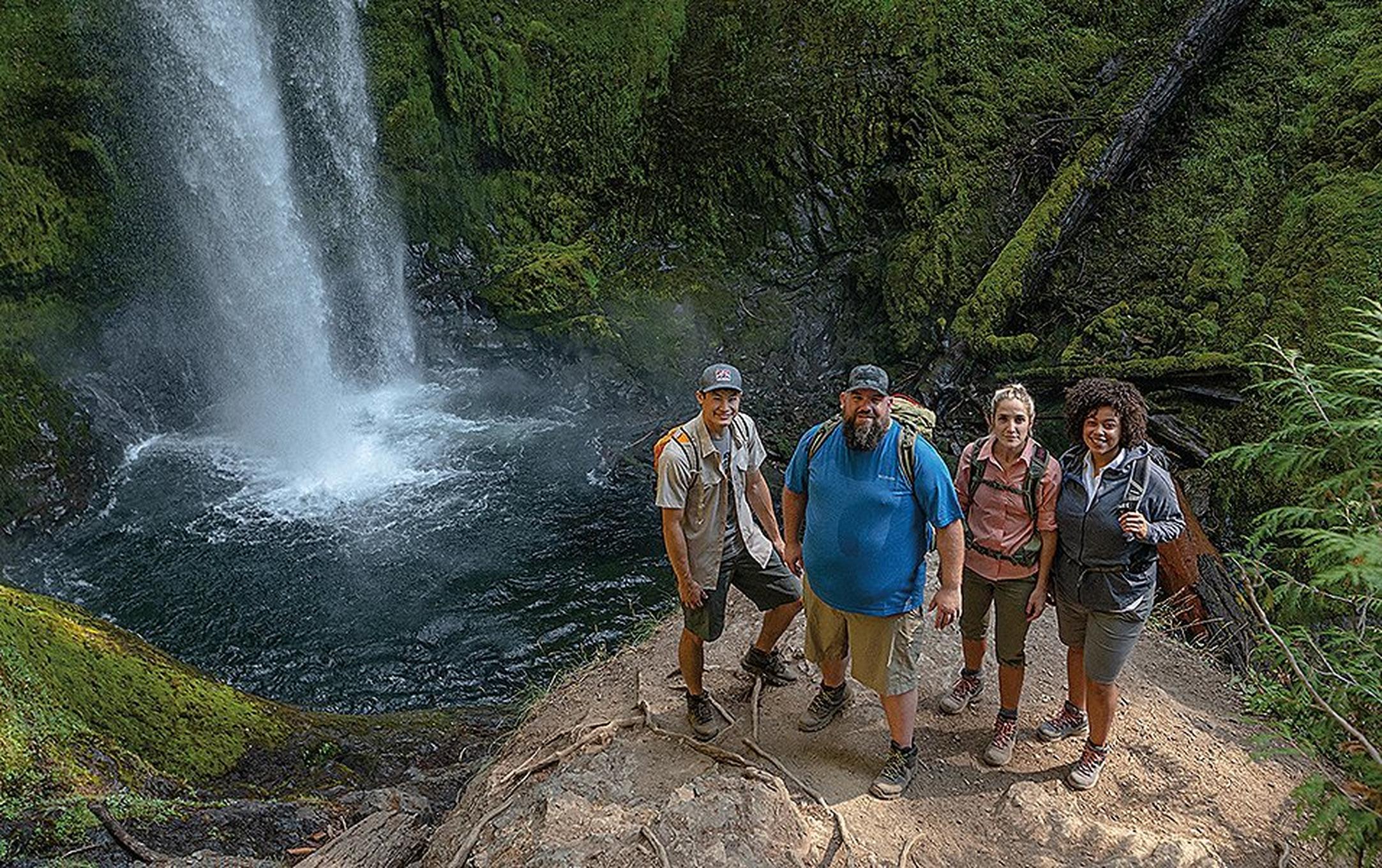 A group of hikers stands in front of a gorgeous waterfall wearing backpacks and smiling at the camera.