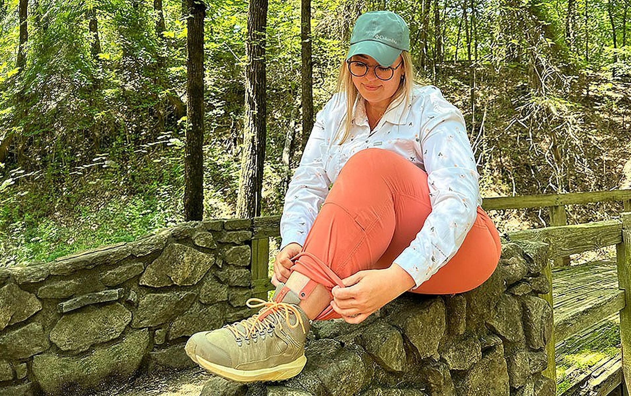 Chelsea McCurdy sits on a pretty stone wall on a hiking trail at Cadron Settlement Park, rolling up the leg of Columbia Sportswear’s Saturday Trail™ Stretch Pants.