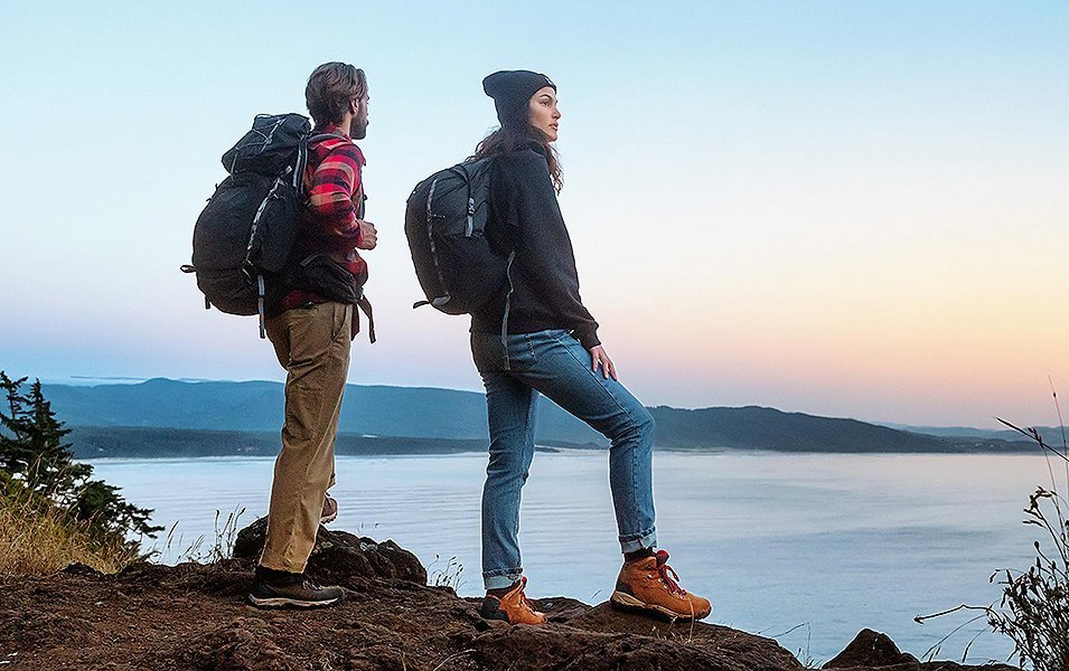 Two hikers stand on a trail overlooking a beautiful body of water at dusk.