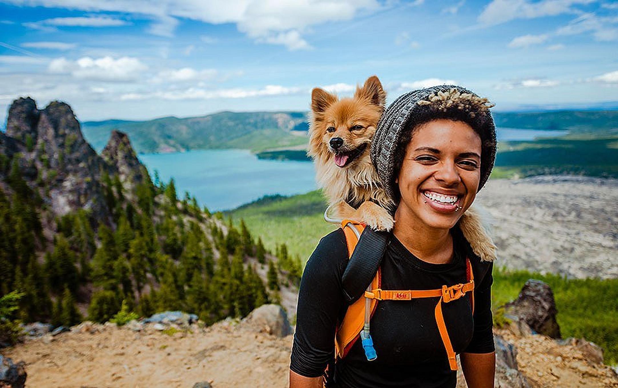 A woman stands atop a beautiful hiking trail with trees and mountains in the background, and her small dog perched on her shoulder.